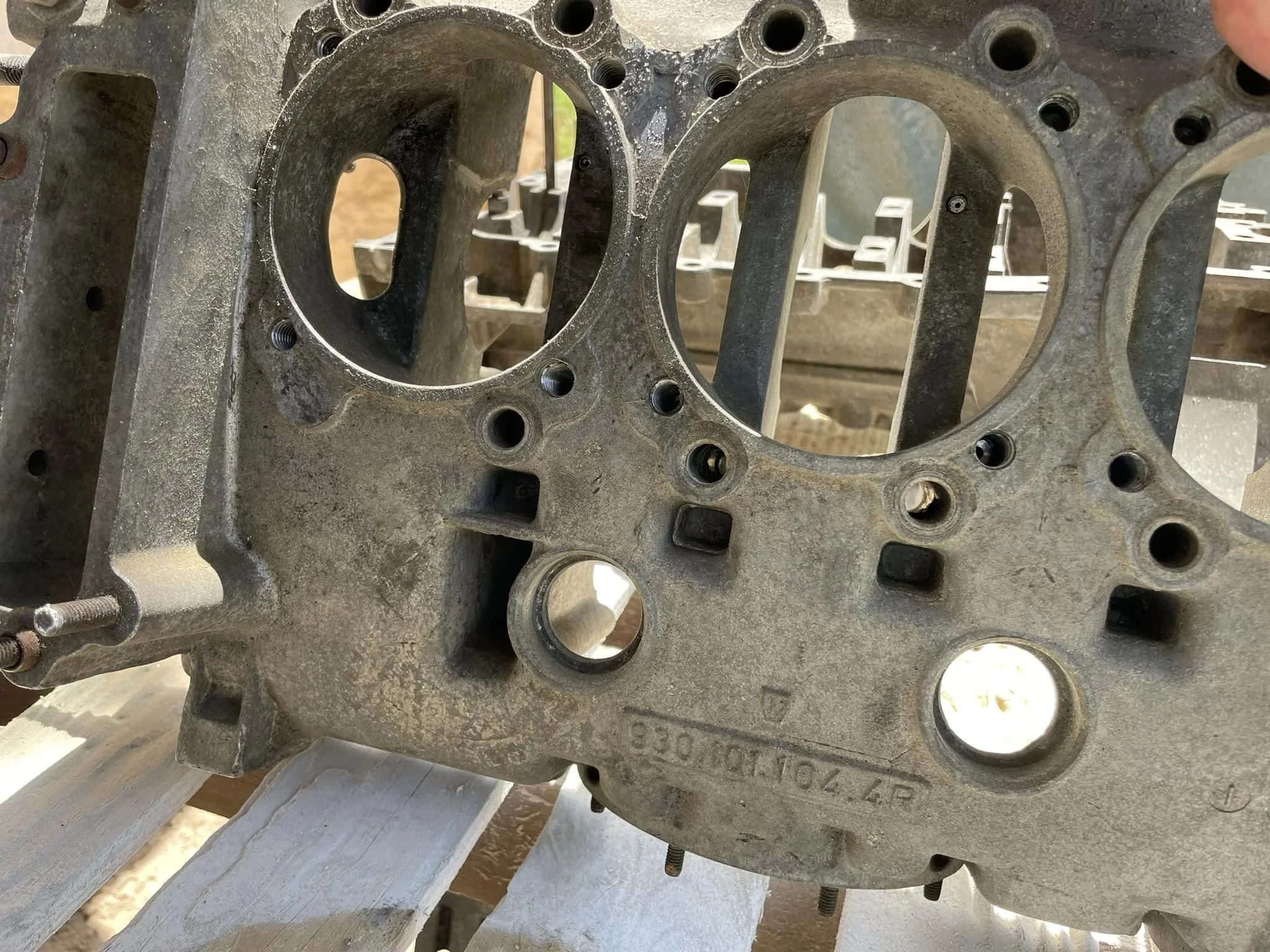 Close-up of a used metal engine block with multiple circular and rectangular cavities, bolts, and a worn surface, resting on a wooden surface outdoors.