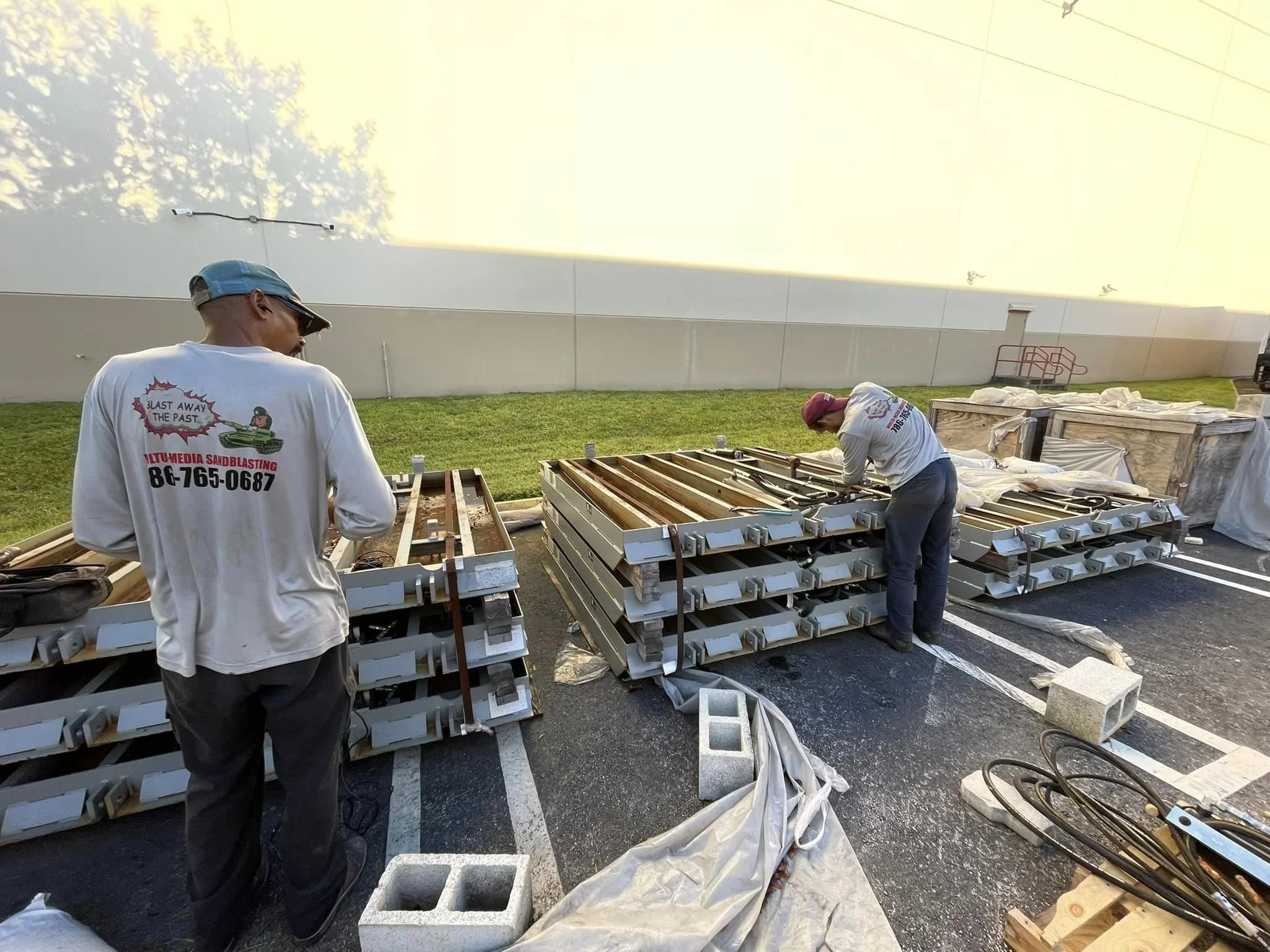 Two workers installing lighting fixtures in a parking lot during daytime, with stacks of metal and boxes around them.