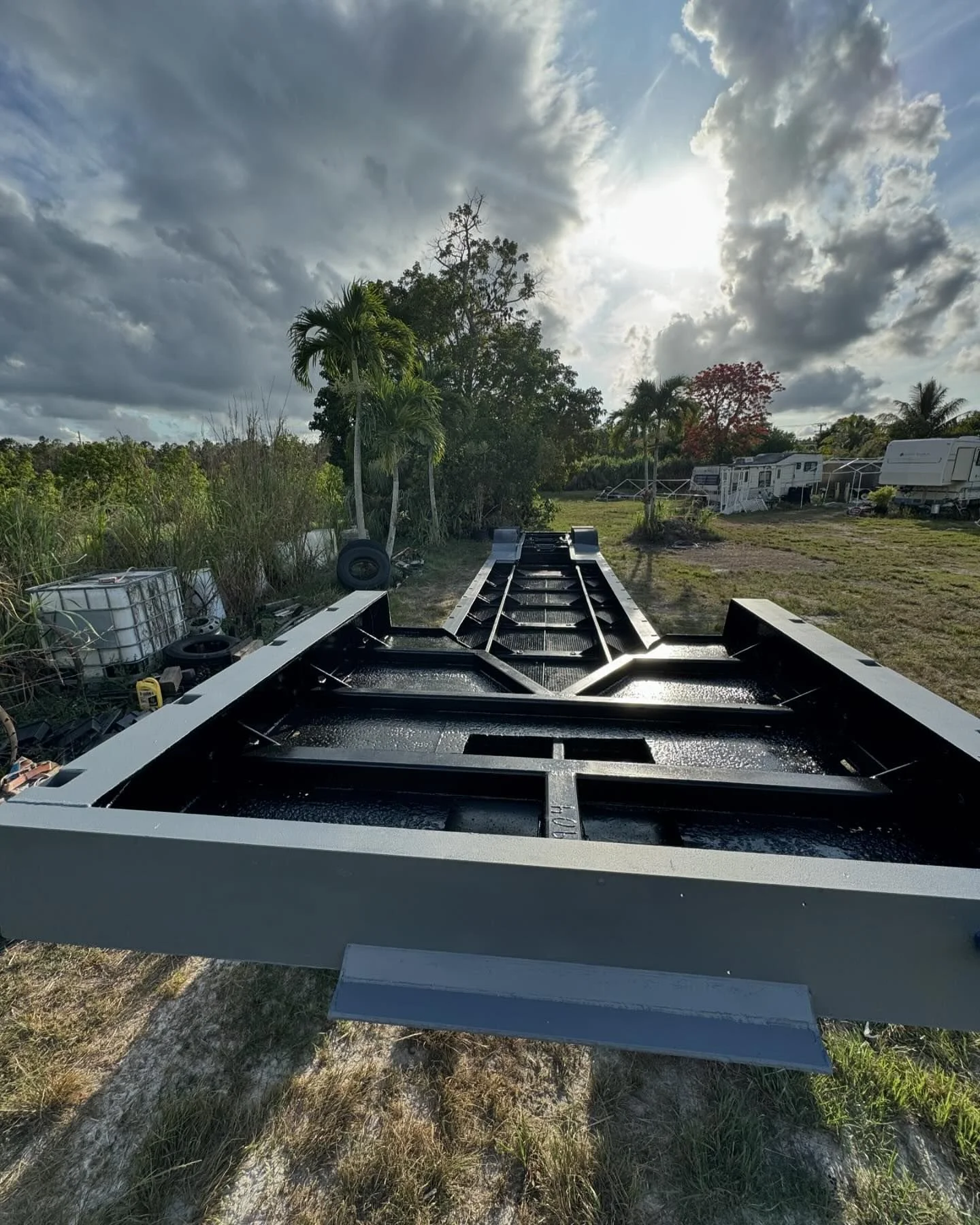 View of a boat hull under construction outdoors with tools and equipment nearby, surrounded by trees and a cloudy sky with the sun partially visible.