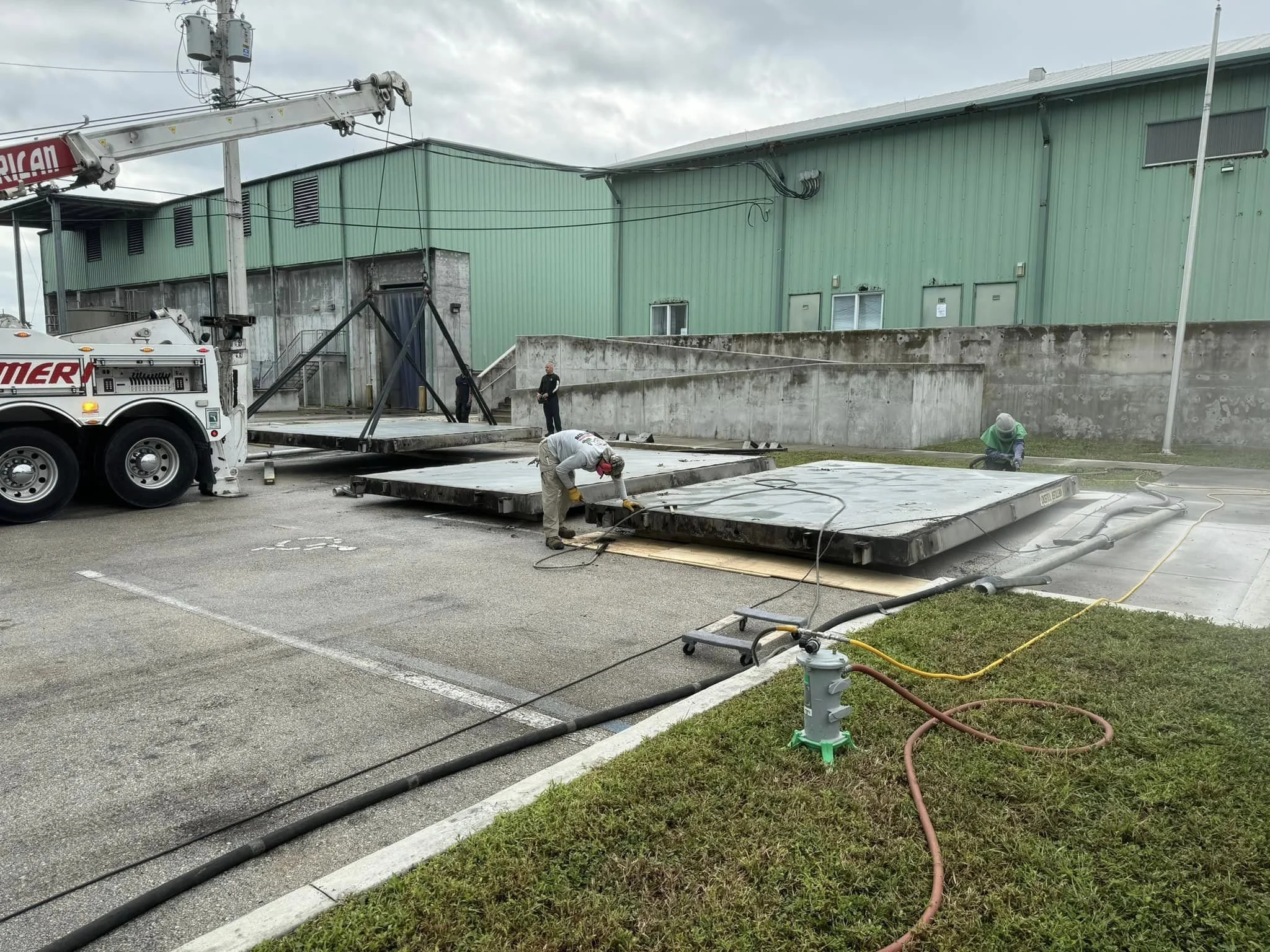 Workers with safety gear are installing large inflated billboard platforms, with a crane truck in a parking lot near a green industrial building.