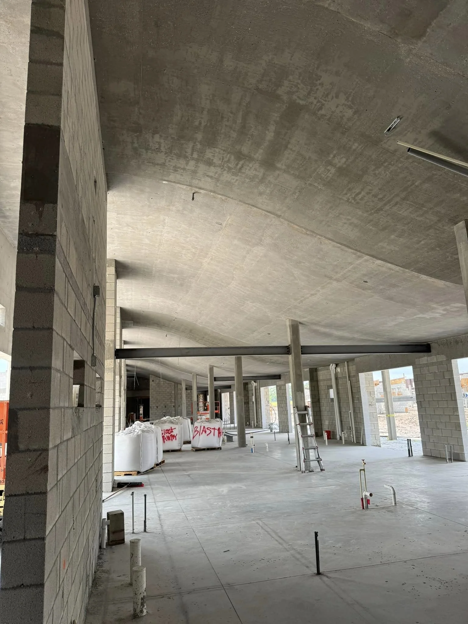 Interior of a building under construction with concrete floors, brick walls, and large open windows. Construction materials and tools are visible, including bags labeled "BLAST" and "CLOSE ARM" and a ladder.