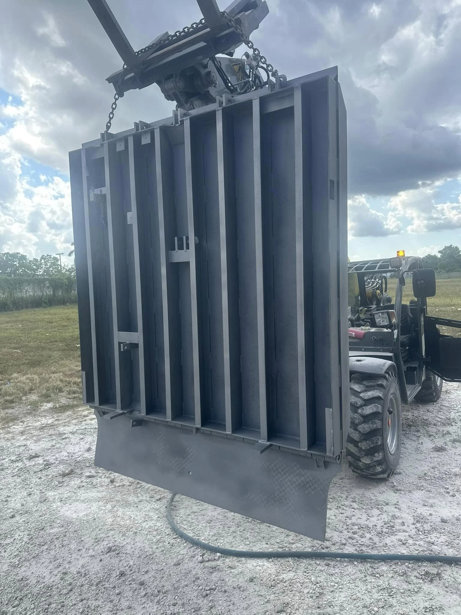 A hydraulic lift holding a large metal cargo container or frame on a forklift in an outdoor setting with a cloudy sky and open field.