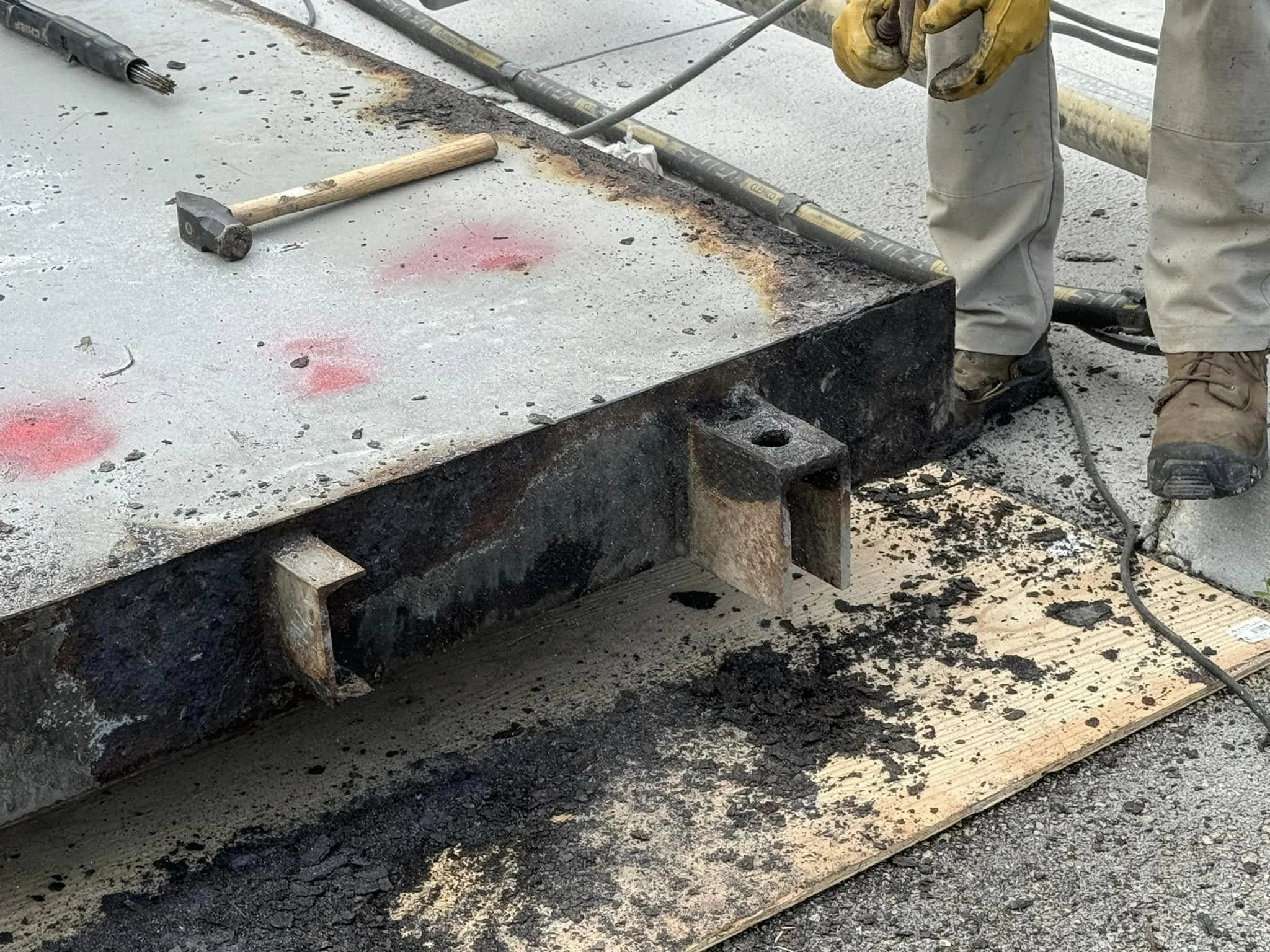 Construction worker cutting or grinding a large metal structure on a construction site, with tools like a hammer placed on the structure and dirt and debris on the ground.