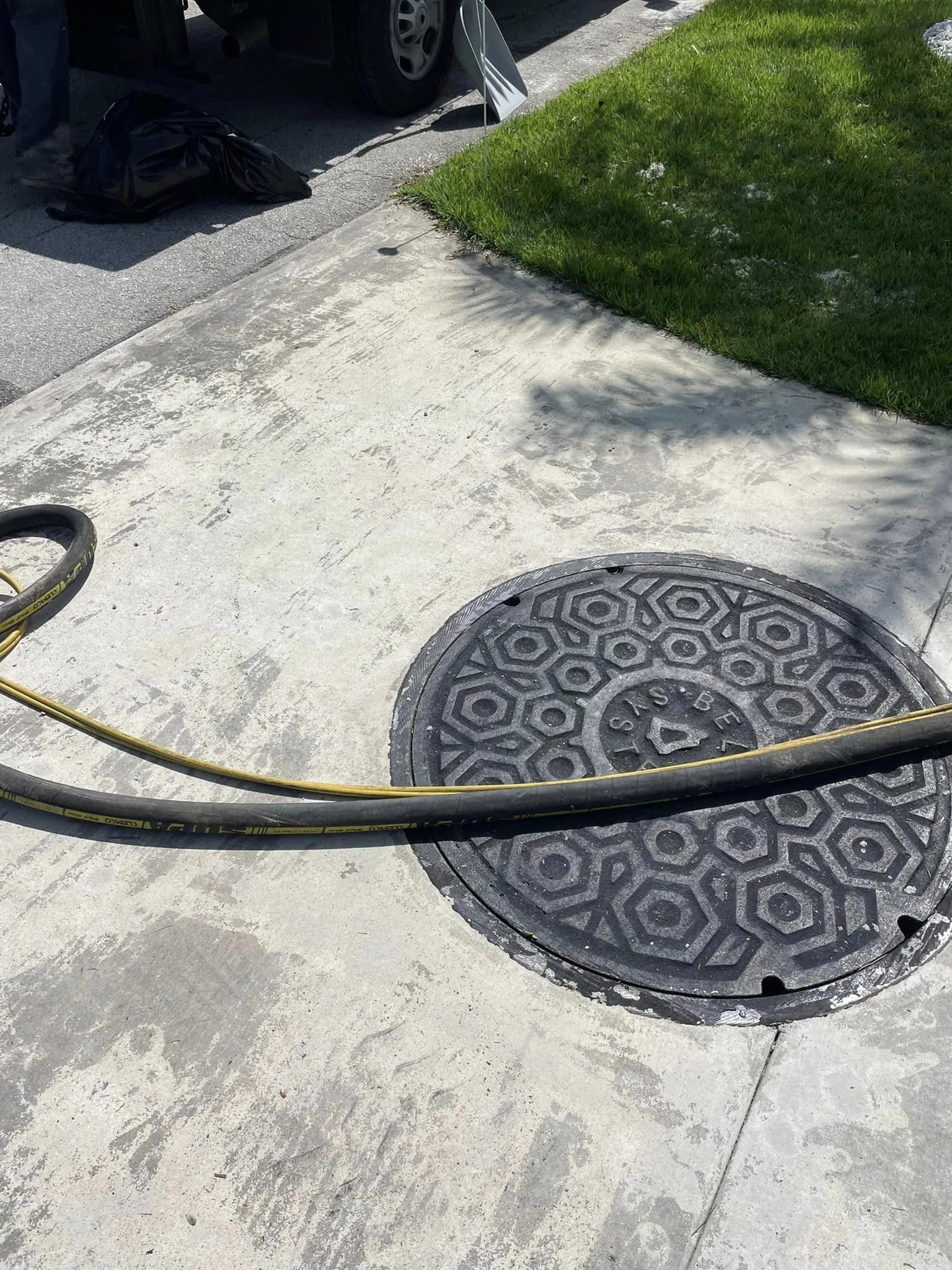 Close-up of a circular storm drain on a concrete sidewalk with a yellow hose partially covering it. In the background, there is a grassy area and building with tools and a trash bag.