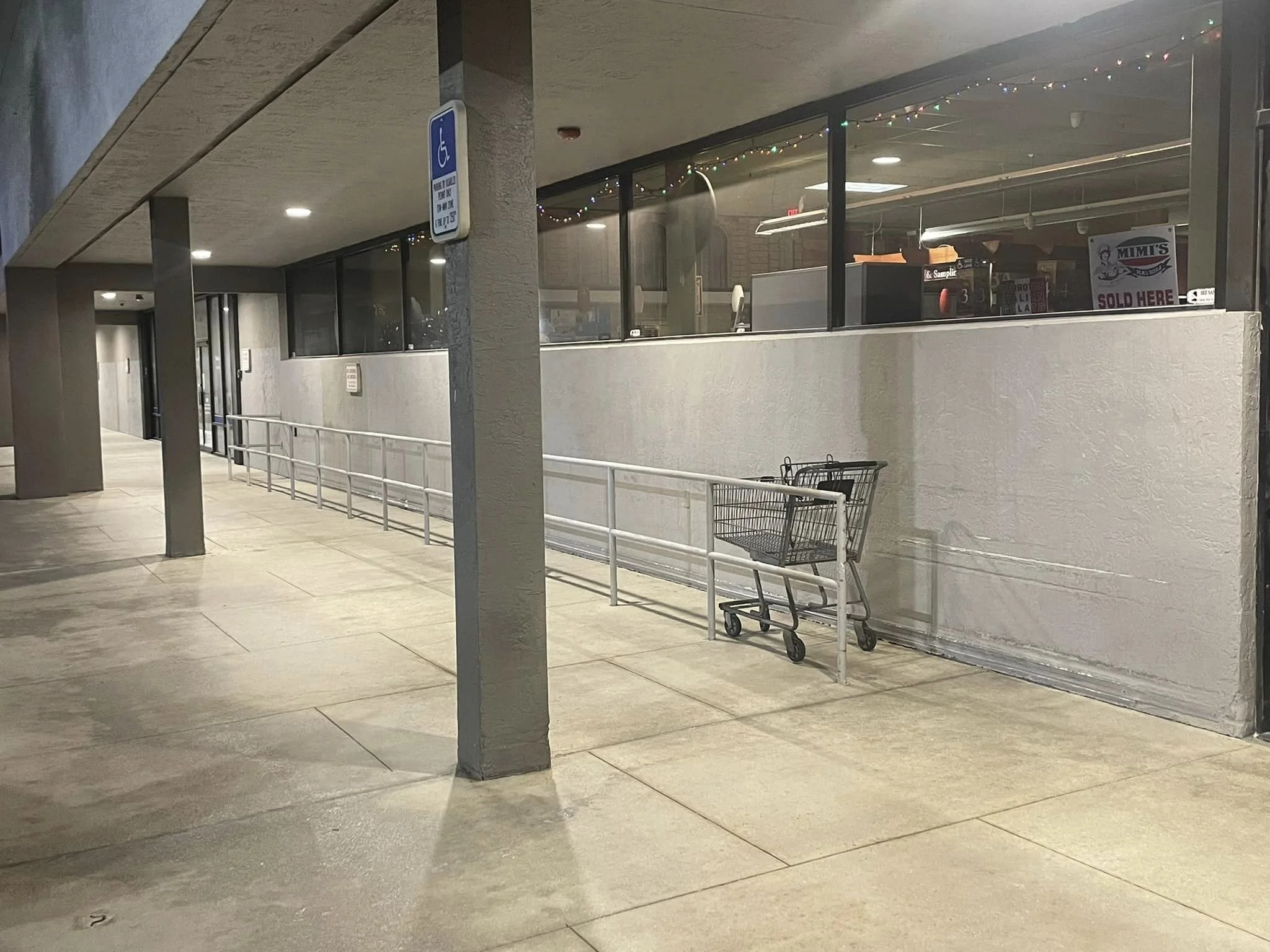 Empty shopping cart positioned next to a parking lot ramp beside a store window decorated with Christmas lights.
