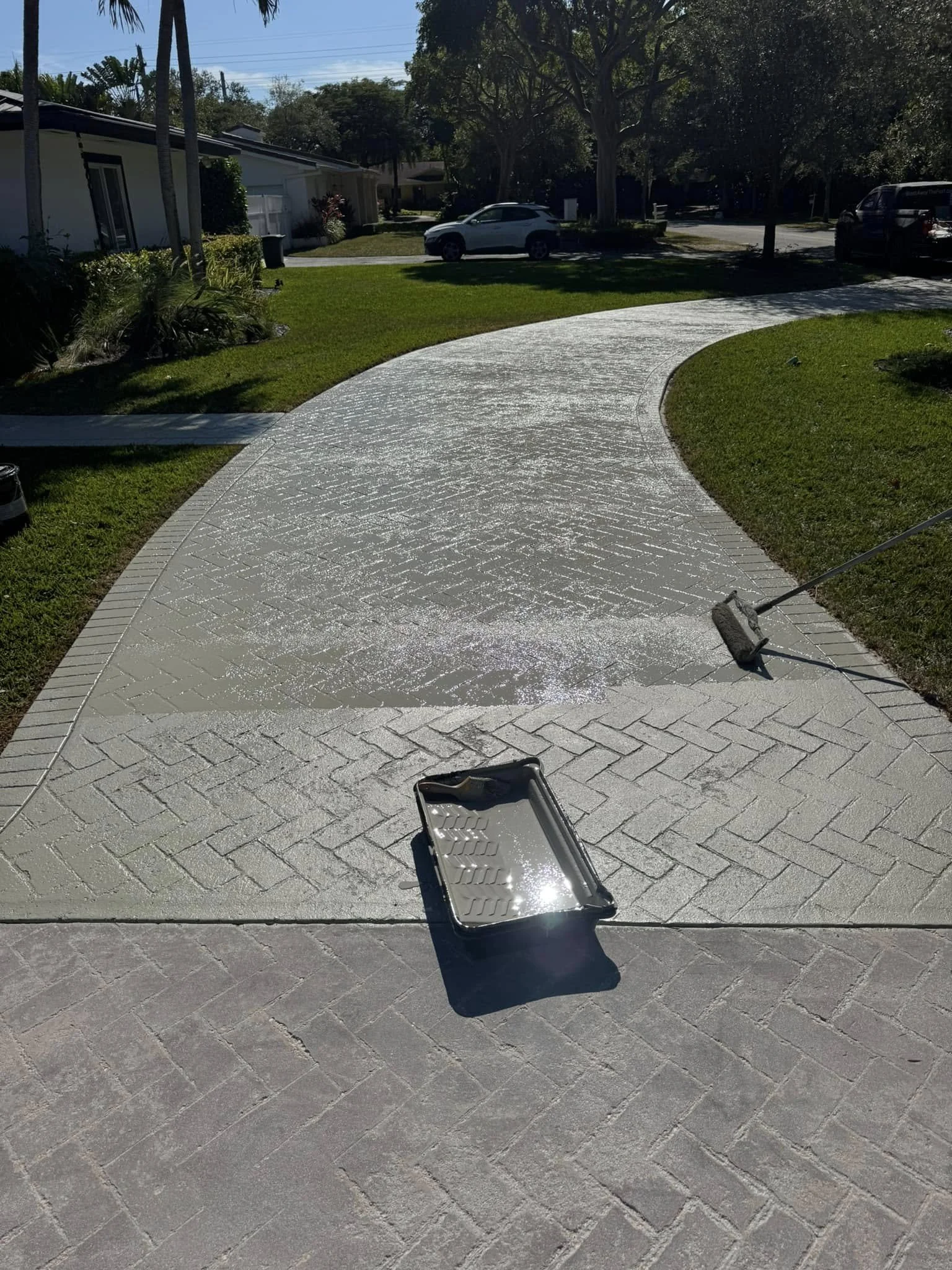 A residential driveway is being pressure washed, with a paint roller attached to a long handle resting on the wet surface. A paint tray filled with water and soap is on the ground nearby. The driveway is made of interlocking bricks and the scene is o