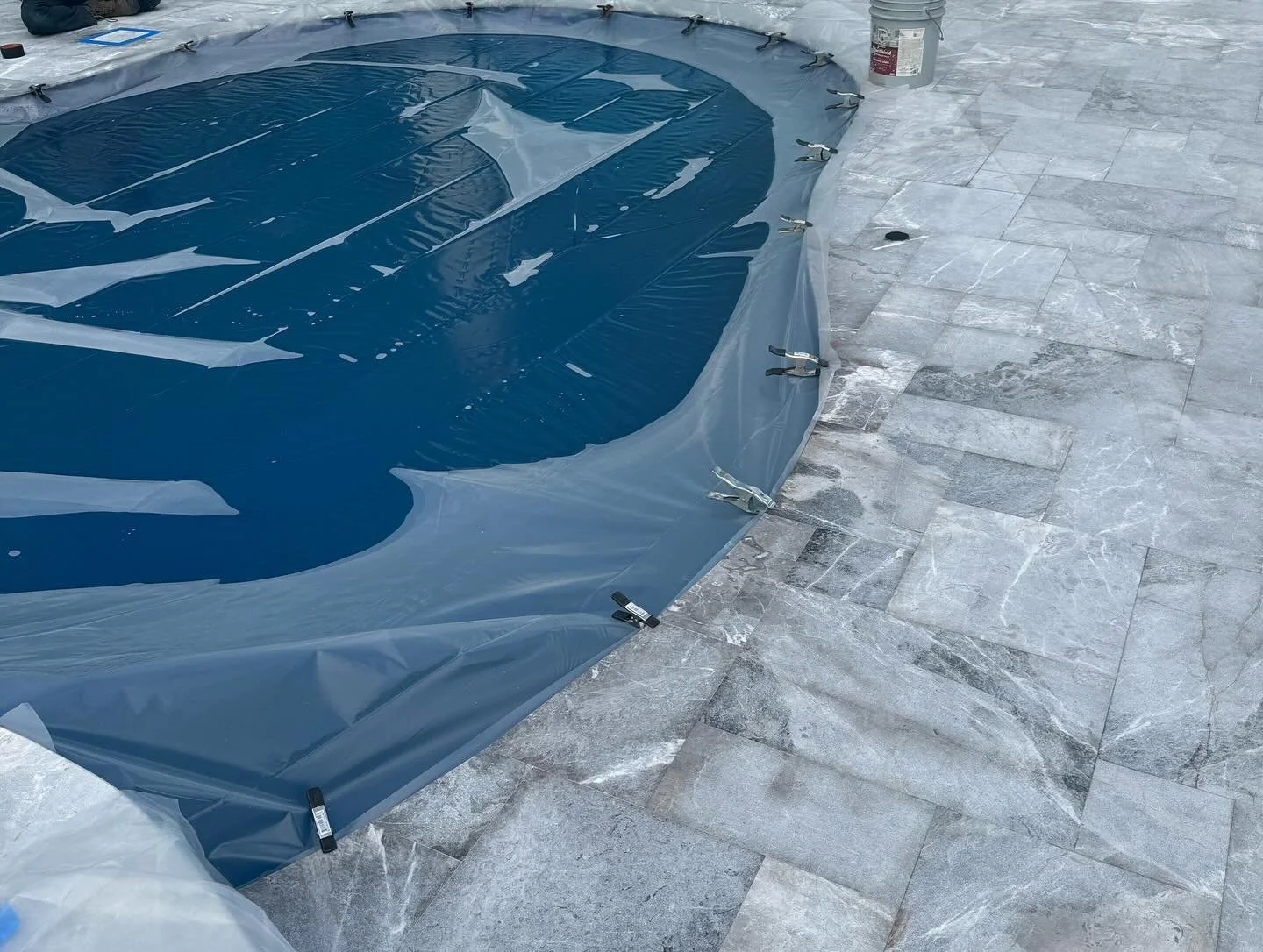Under construction or renovation of a swimming pool, with blue plastic cover and gray stone tiles surrounding it.