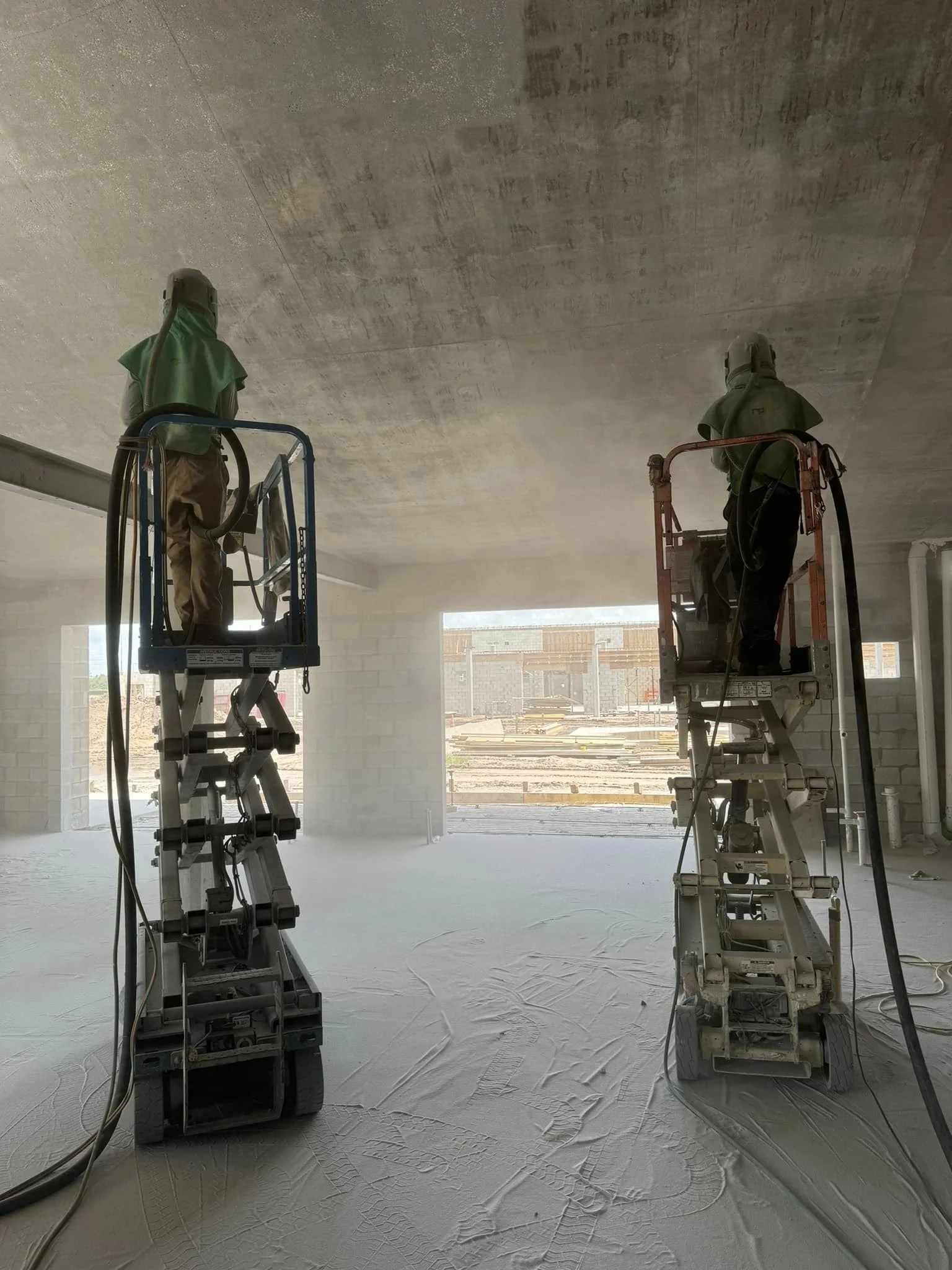 Two construction workers in green safety gear operating lift platforms inside a building under construction.