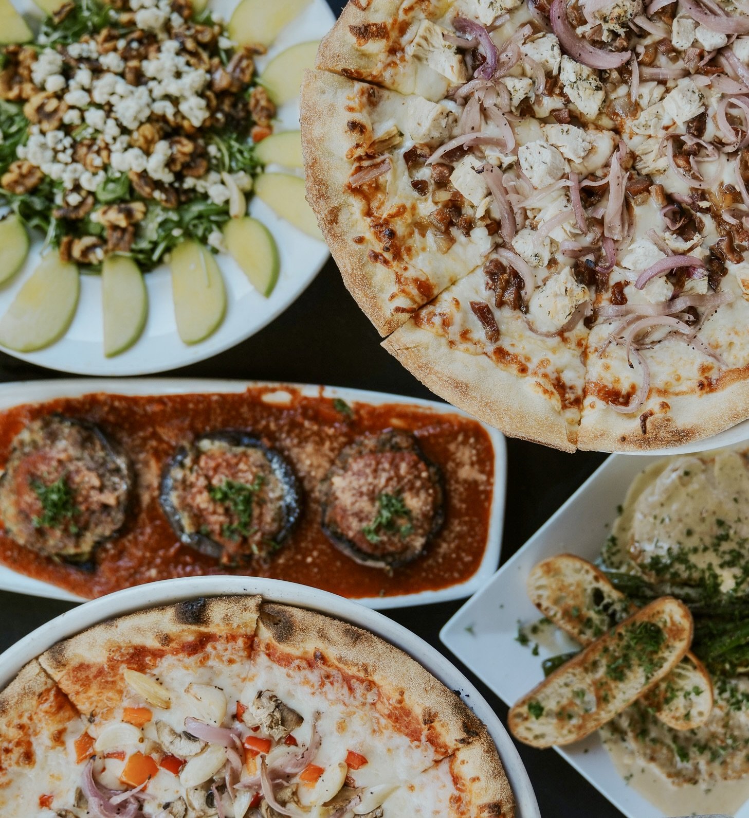 A variety of Italian dishes including pepperoni pizza, chicken pizza, stuffed eggplant with tomato sauce, and garlic bread with herbs.