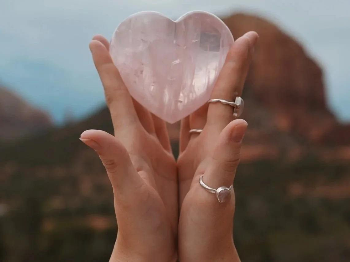 Cupped hands hold a heart-shaped gemstone up toward the red rocks of Sedona.