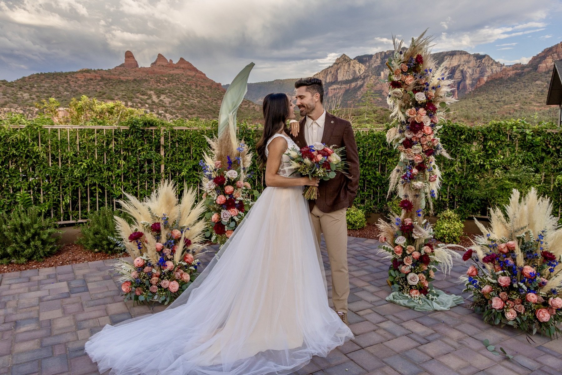 A bride and groom standing together outdoors, wedding ceremony backdrop with floral arrangements, mountainous landscape in the distance, cloudy sky.