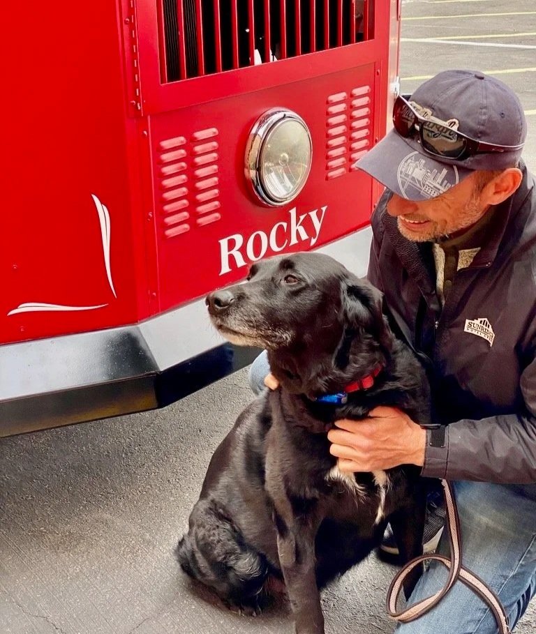 A man pets a black dog in front of the Sedona Trolley.