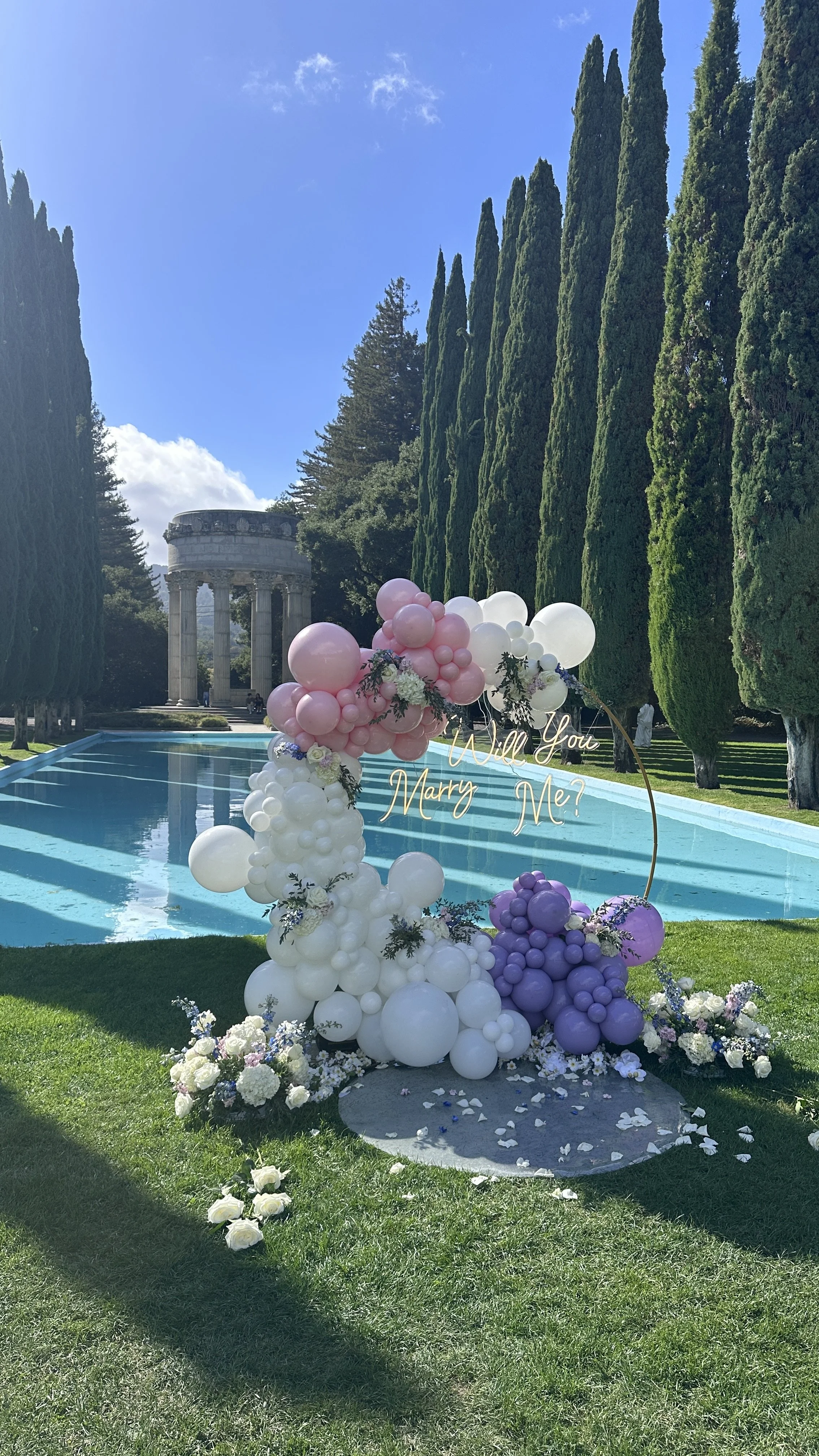 Will You Marry Me lettering on a mirrored backdrop framed by pastel balloons and white flowers at Filoli Gardens.