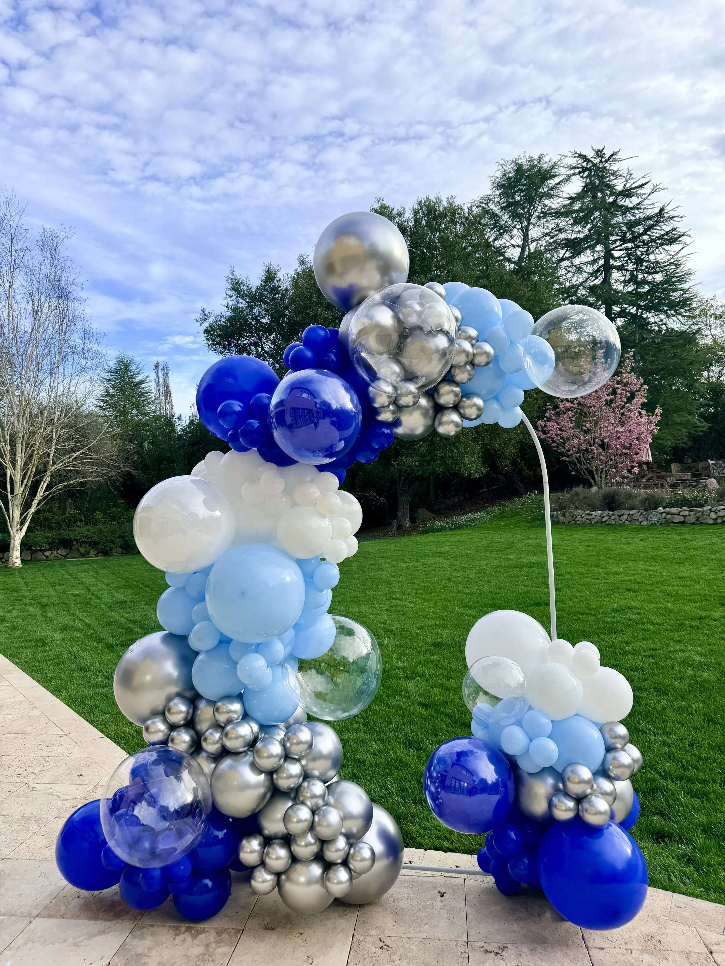 Reflective silver and clear balloons among blue and white clusters on an outdoor graduation garland in Atherton.