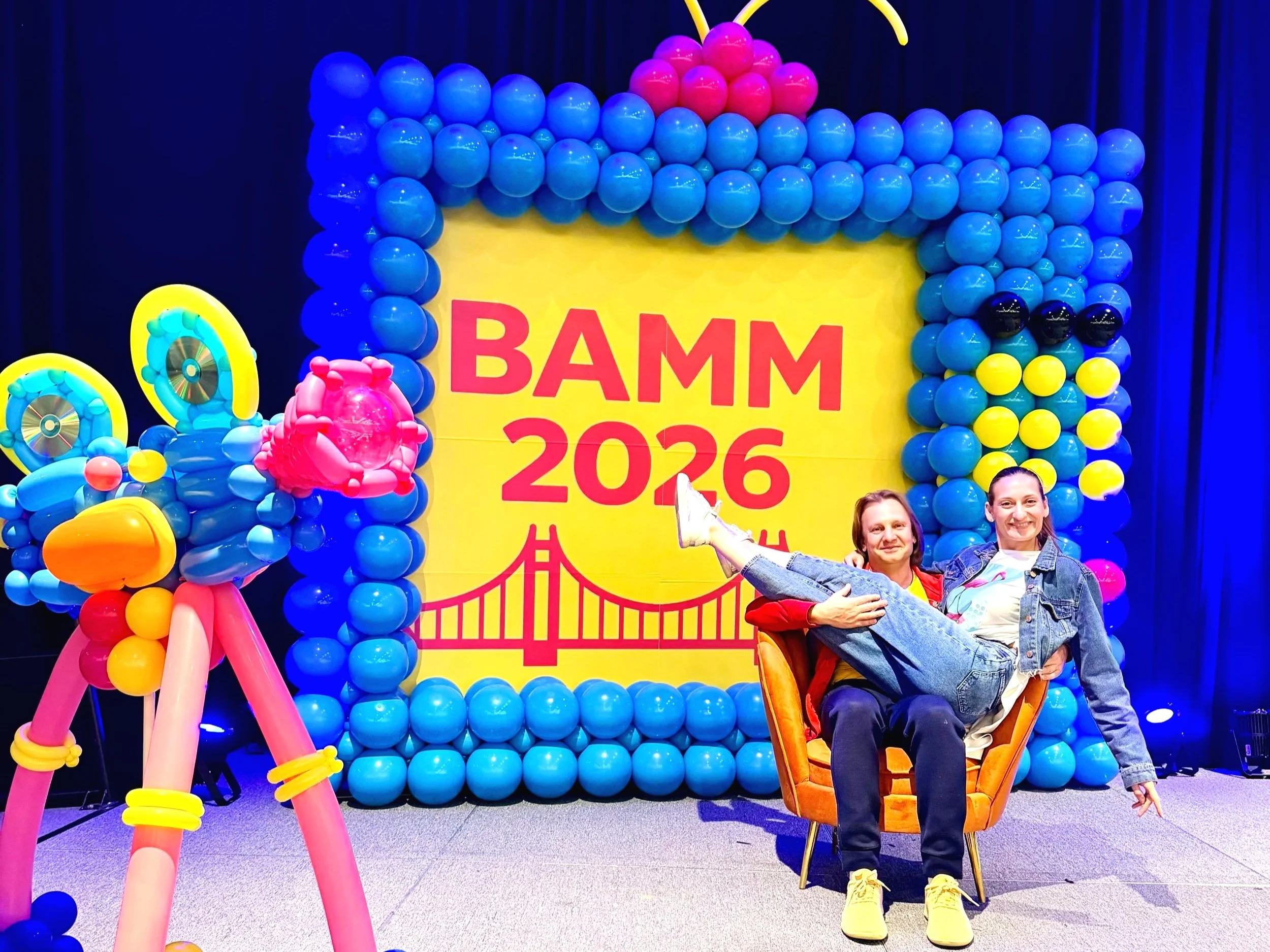 Two women sit happily on an orange chair in front of a vibrant balloon display at Bamm 2026 event. The backdrop features a large yellow sign with pink text and a bridge illustration, surrounded by a colorful balloon arch and sculptures in pink, yellow, and blue.