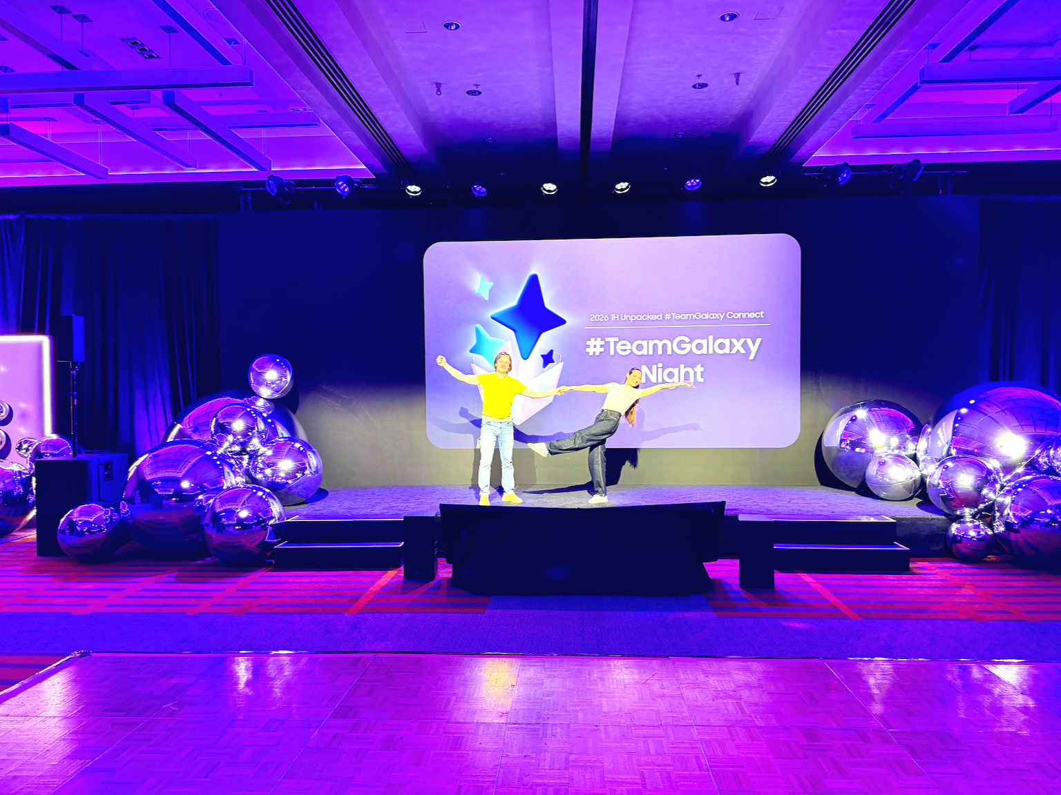 Stage with two performers, one standing and one leaning, during #TeamGalaxy Night event with purple lighting, large metallic balloons on both sides, and a screen displaying event details in the background.