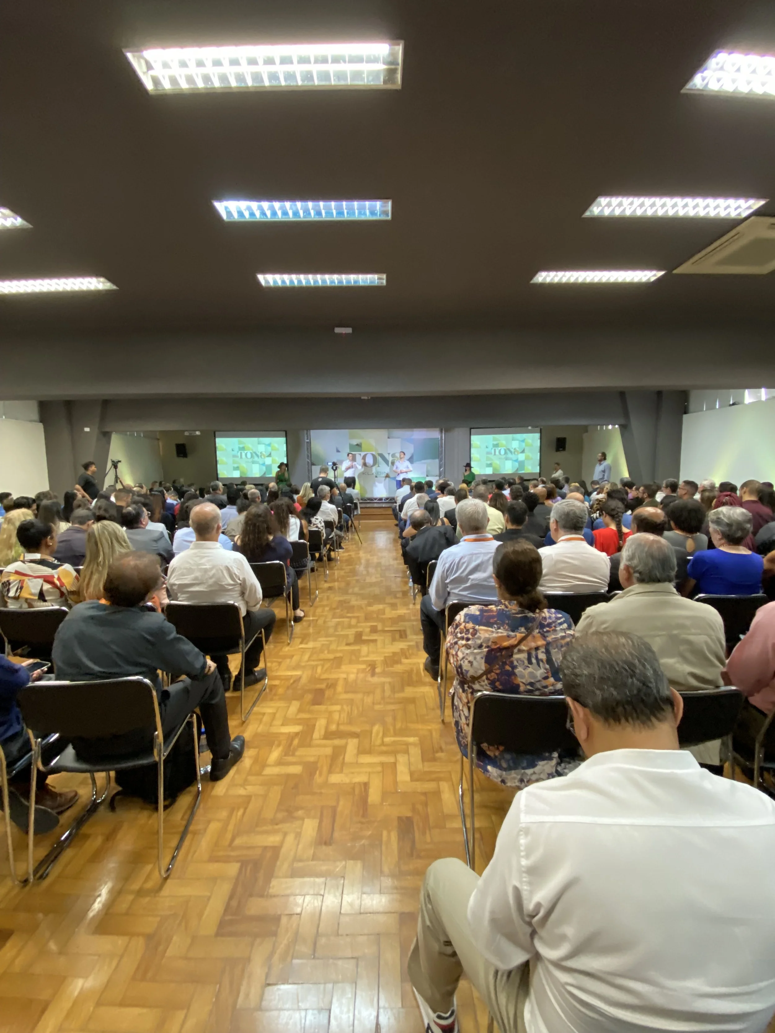 Uma grande sala de conferência cheia de pessoas assistindo a uma apresentação em um palco na frente, com várias telas de projeção.