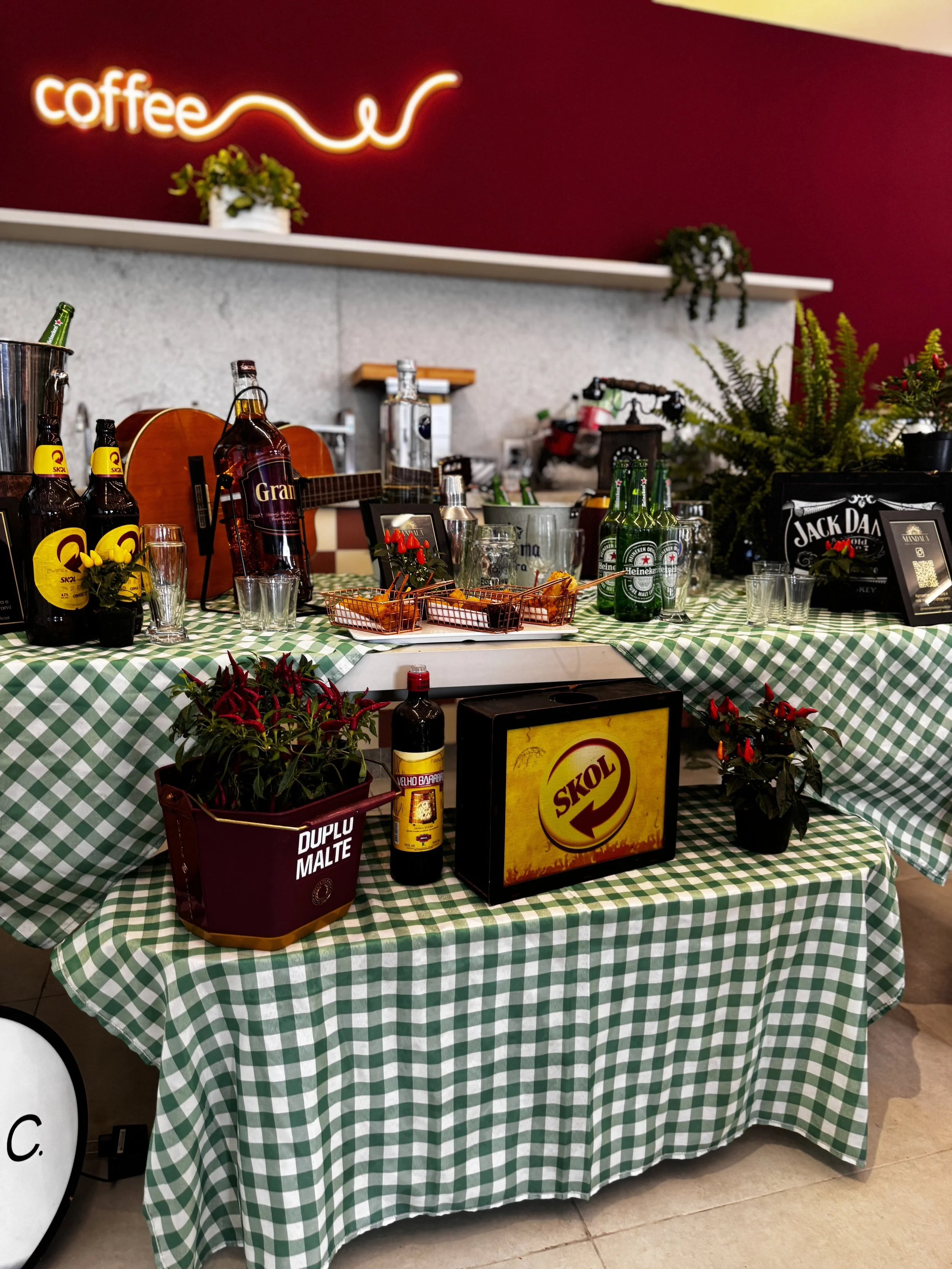 Mesa decorada com tema do Skol, com plantas, garrafas de cerveja, copos, uma guitarra e itens de assinatura da marca, com toalhas verdes xadrez e fundo com parede vermelha e iluminação de neon 'coffee'.