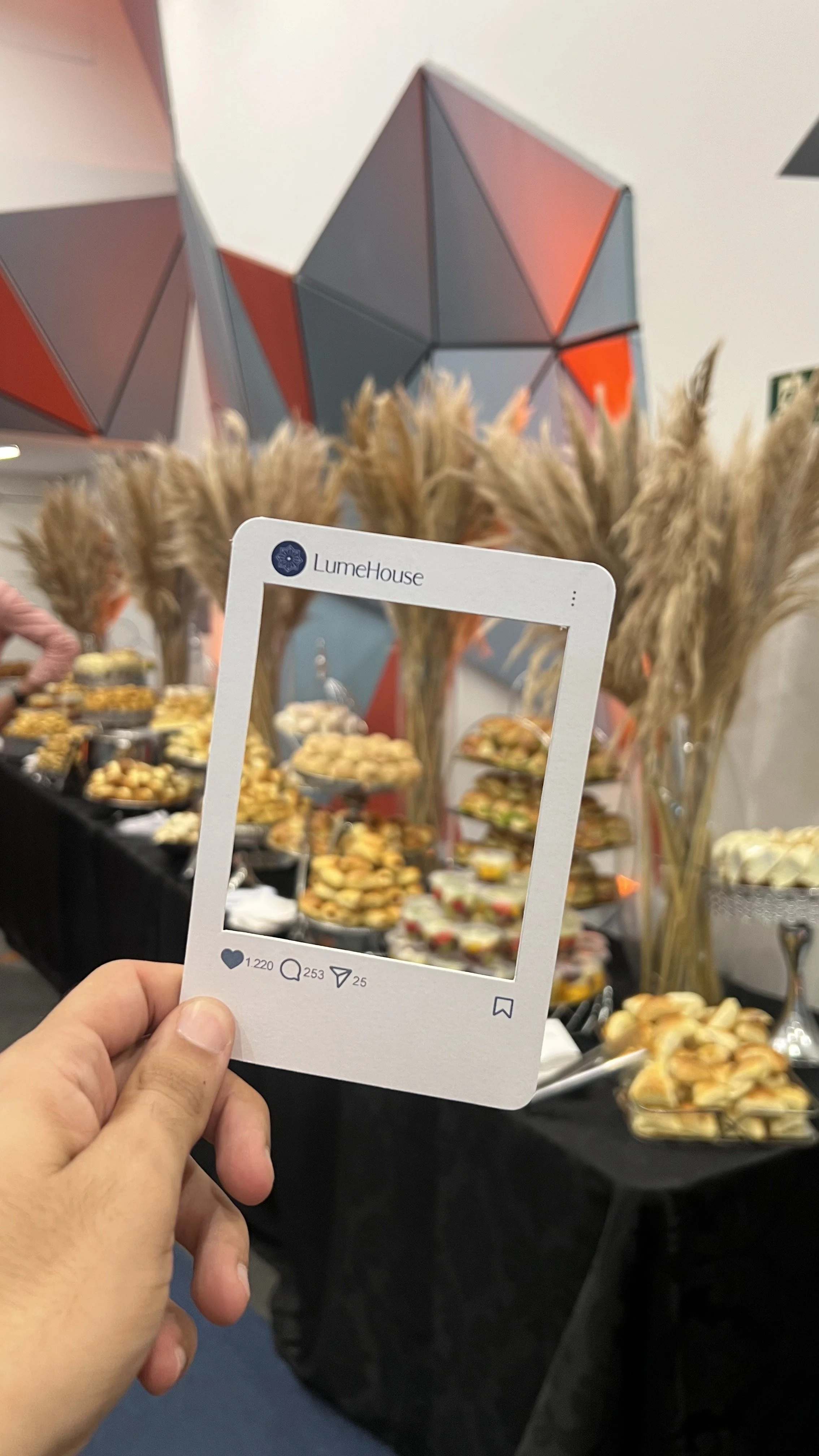 Mão segurando uma moldura que imita uma postagem do Instagram, com o logo 'LumeHouse'. Ao fundo, um buffet de doces e aperitivos, decorado com pratos e flores secas, com uma parede com uma instalação geométrica de cores vermelha, cinza e azul.
