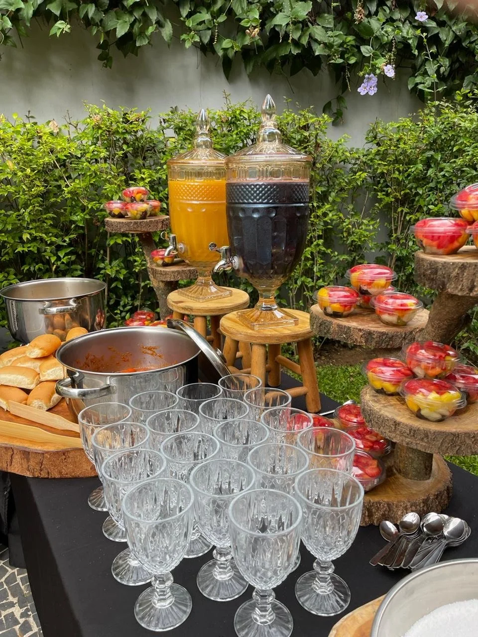 Mesa de festa com bebidas, copos de vidro, doces e frutas decoradas em caixinhas plásticas transparentes, ao ar livre com plantas ao fundo.