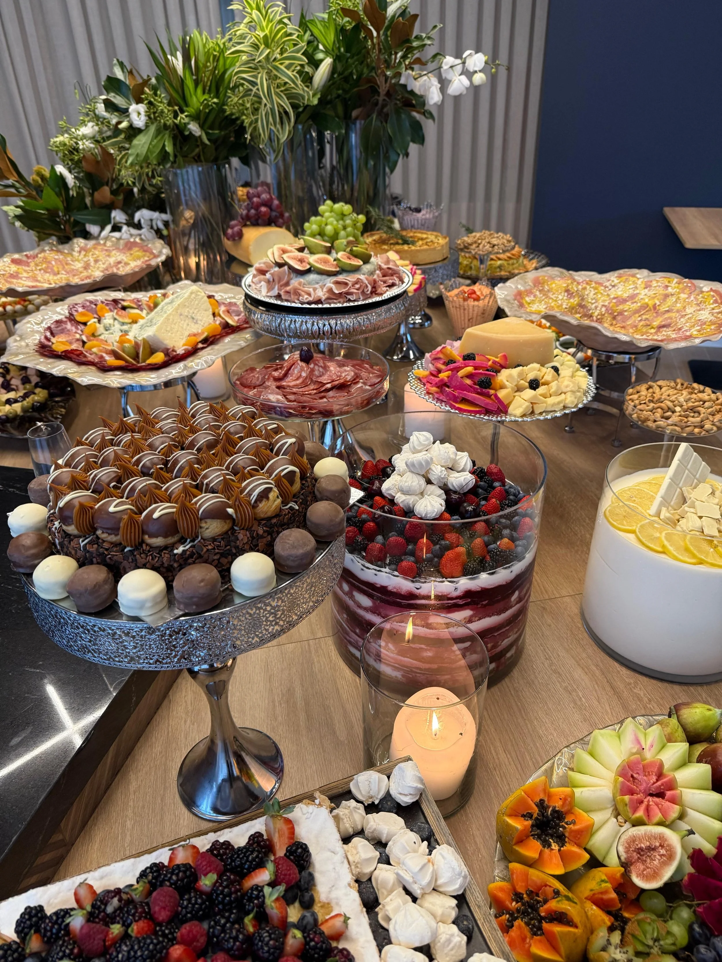 Mesa repleta de doces e frutas, incluindo bolo decorado com frutas vermelhas, tortas, macaron, bolo de chantilly com limão, frutas como melão, figo, uva e morango, além de decorações de flores e velas.