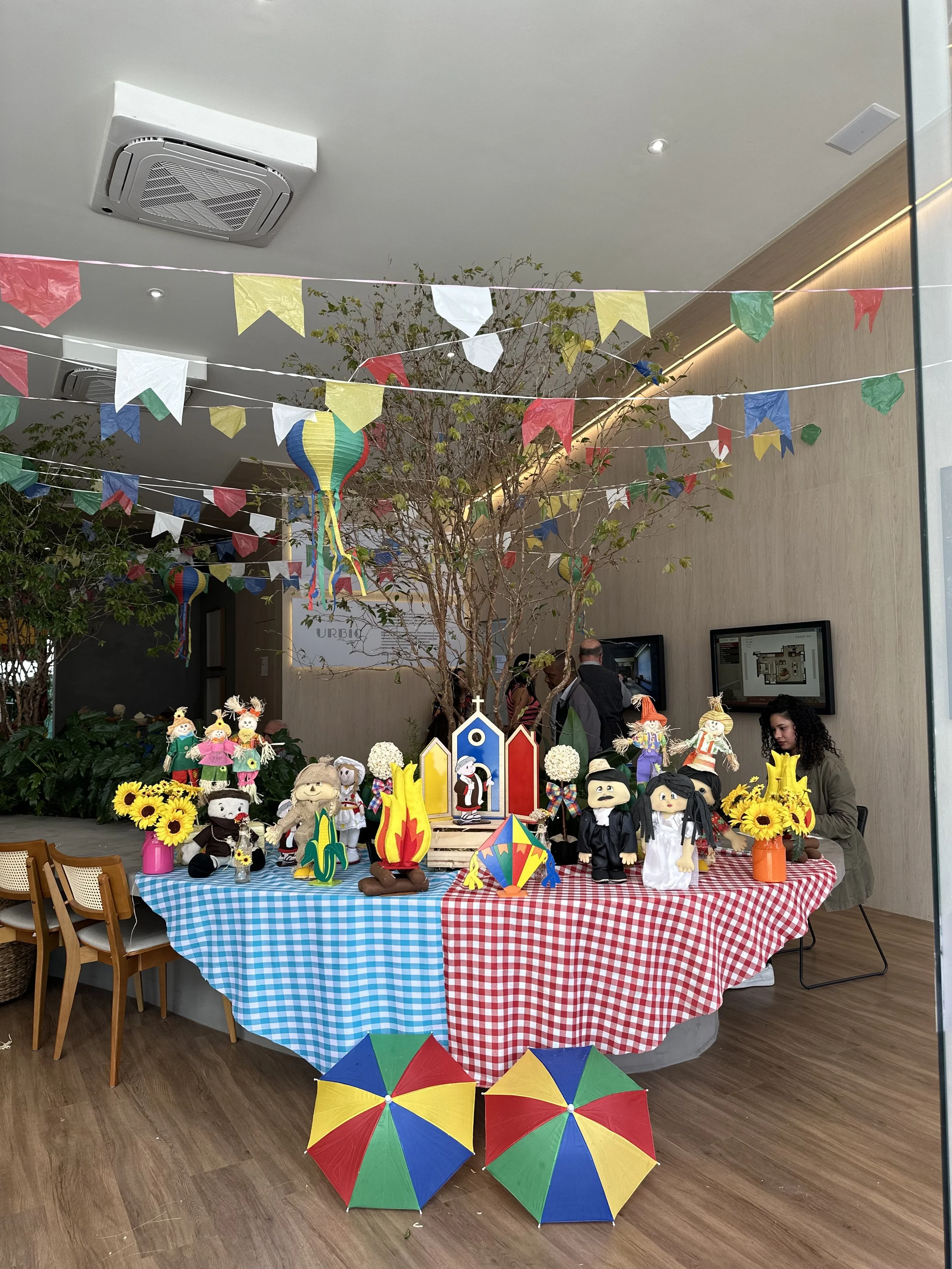 Decoração de festa com elementos tradicionais do Brasil, incluindo bonecos representando personagens típicos, enfeites coloridos, bandeirinhas de papel e guarda-chuvas decorativos. Mesa decorada com toalha de chita colorida, flores de girassol e obje