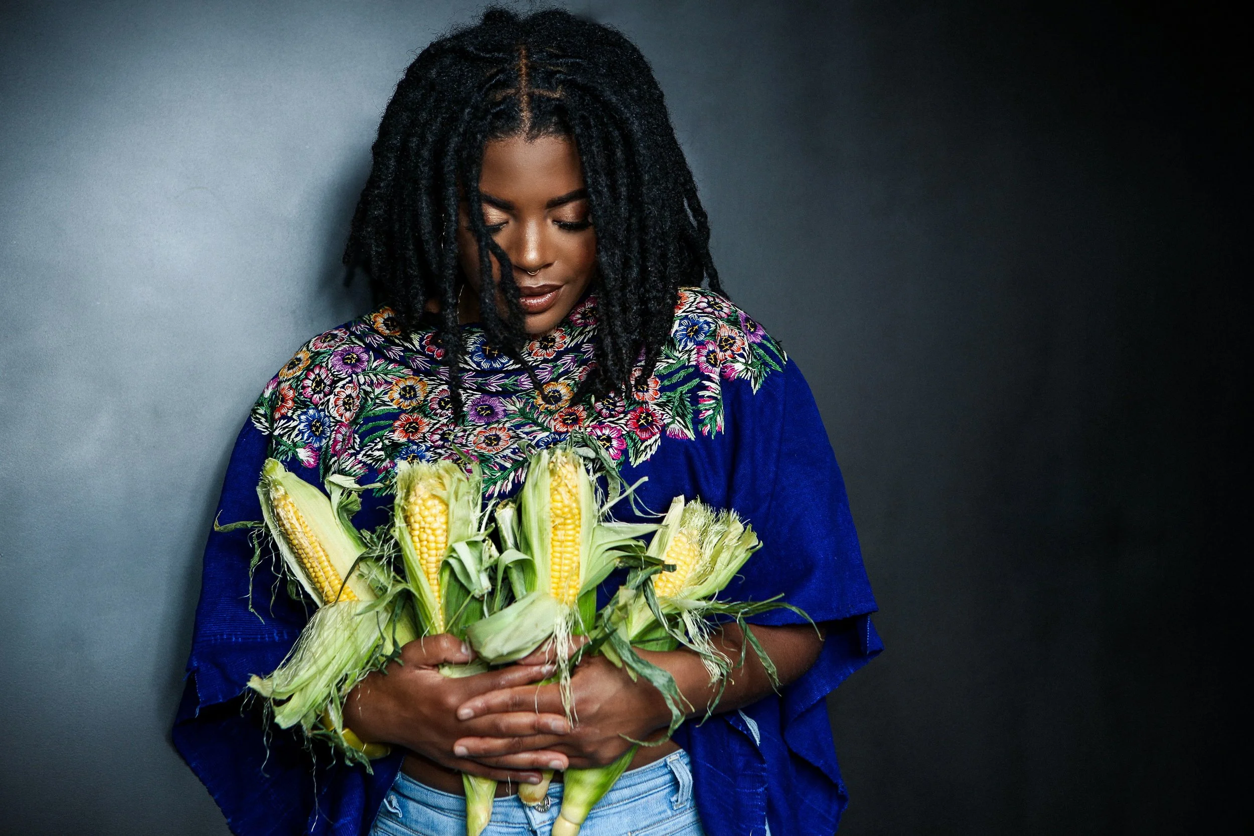 A woman with dreadlocks looks down at a bouquet of corn cobs she is holding.
