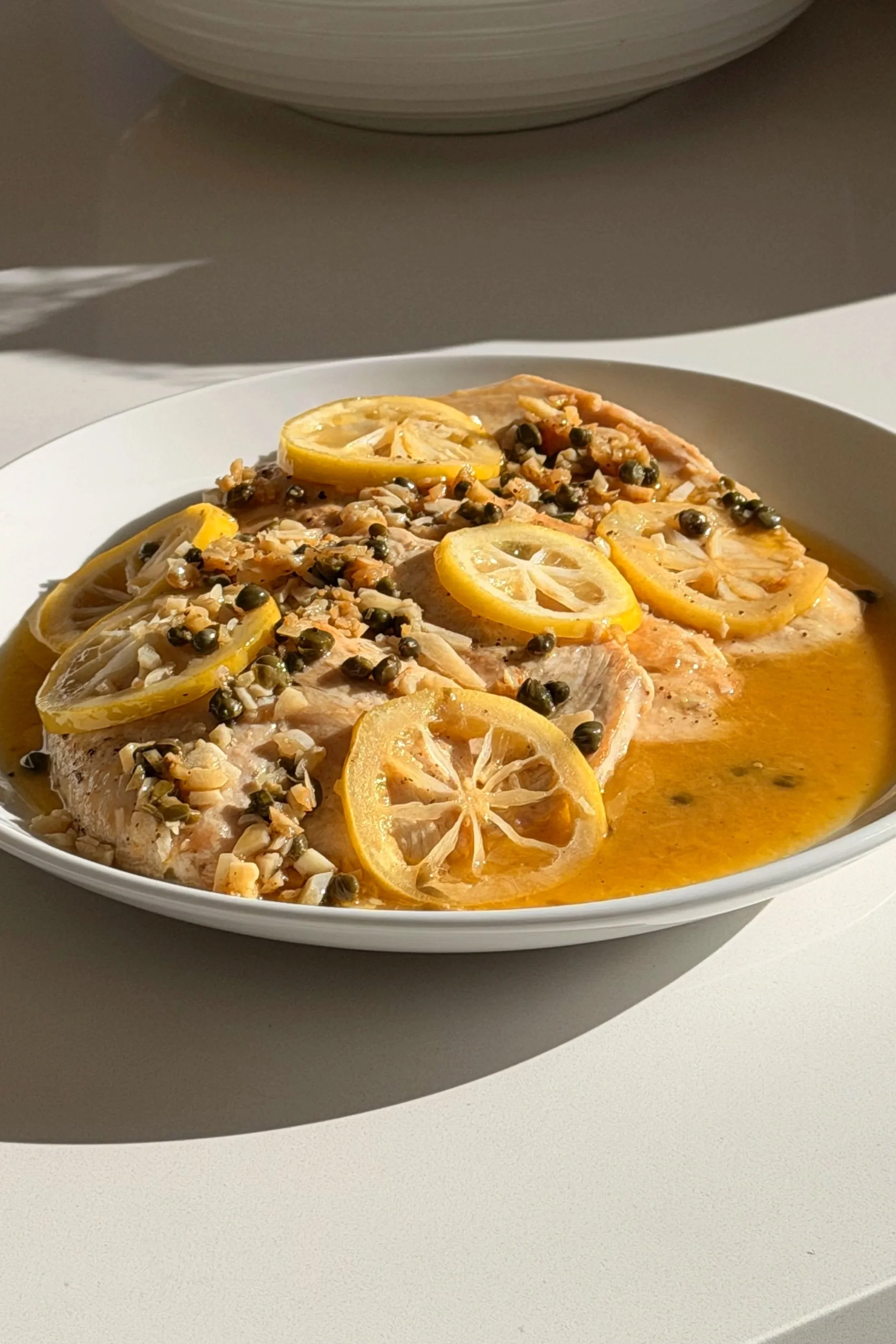 A white plate with cooked fish topped with lemon slices, chopped nuts, and capers, served with a lemon butter sauce.