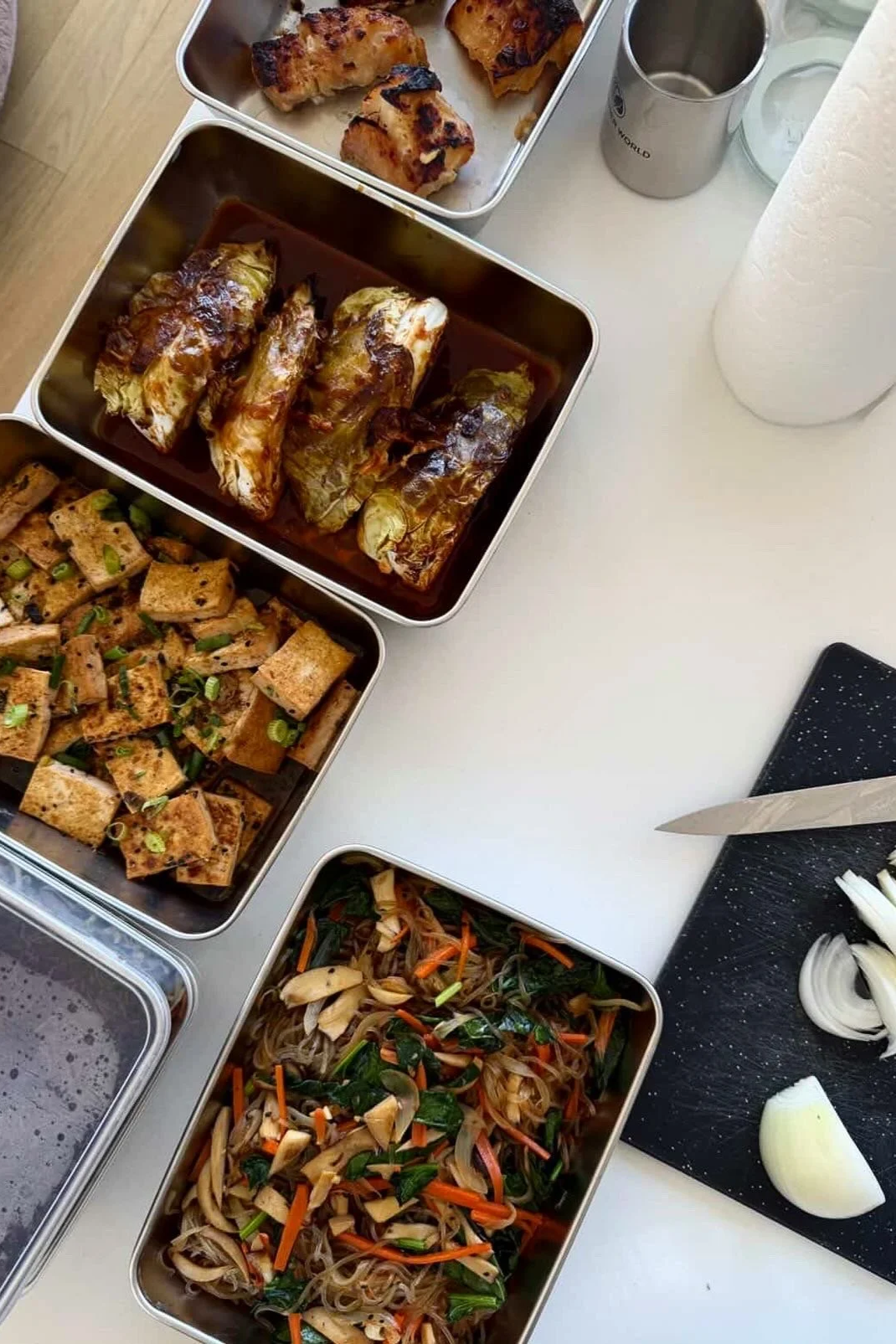 A variety of Asian-inspired dishes in metal containers on a white table, including spring rolls, tofu, stir-fried vegetables, and grilled chicken, with a black cutting board with chopped onions and a knife nearby.
