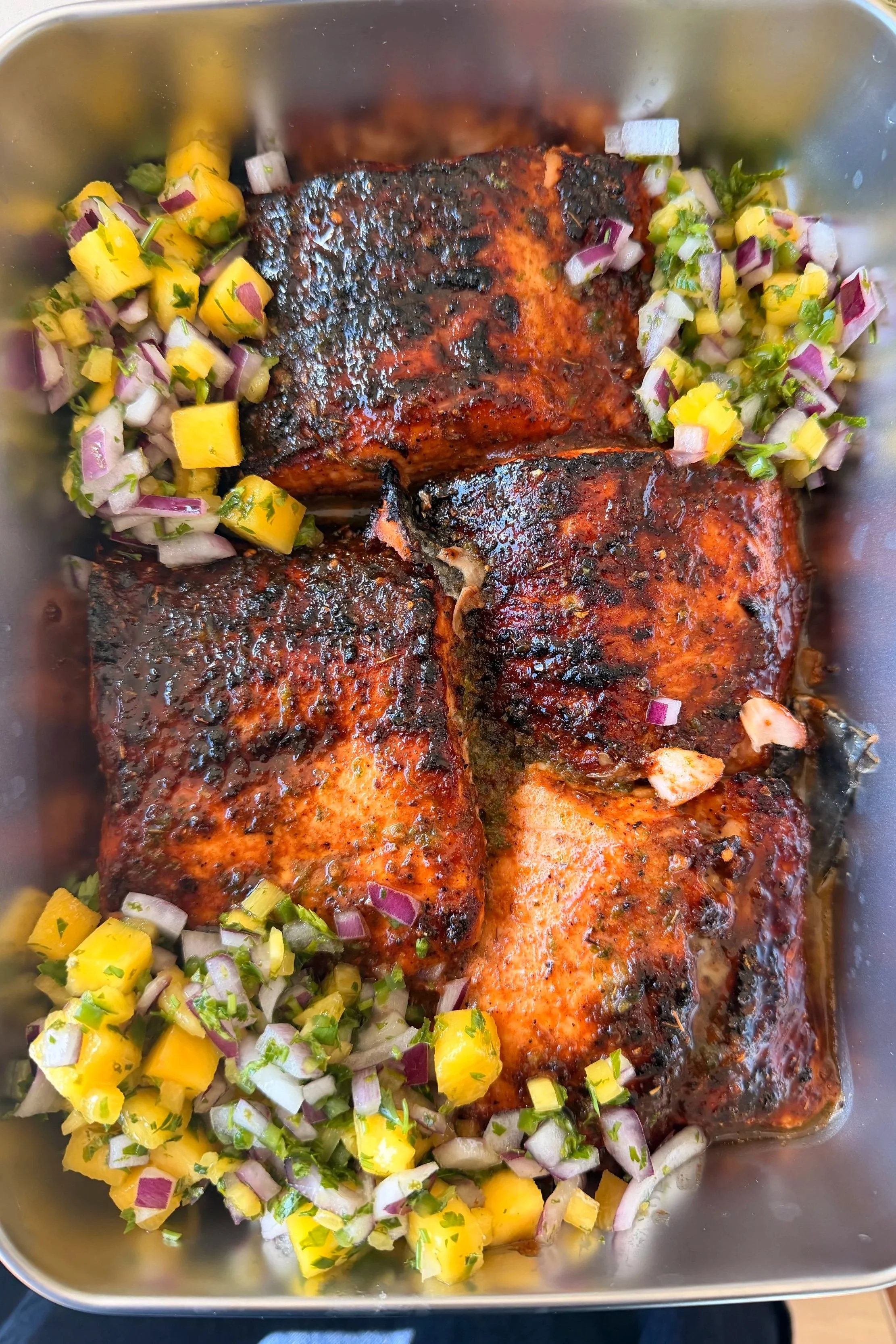 Baked salmon fillets with a dark glaze, garnished with chopped onions, mango, and cilantro in a metal tray.