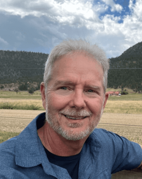 A smiling older man with gray hair and a beard taking a selfie outdoors with a field, trees, hills, and a partly cloudy sky in the background.