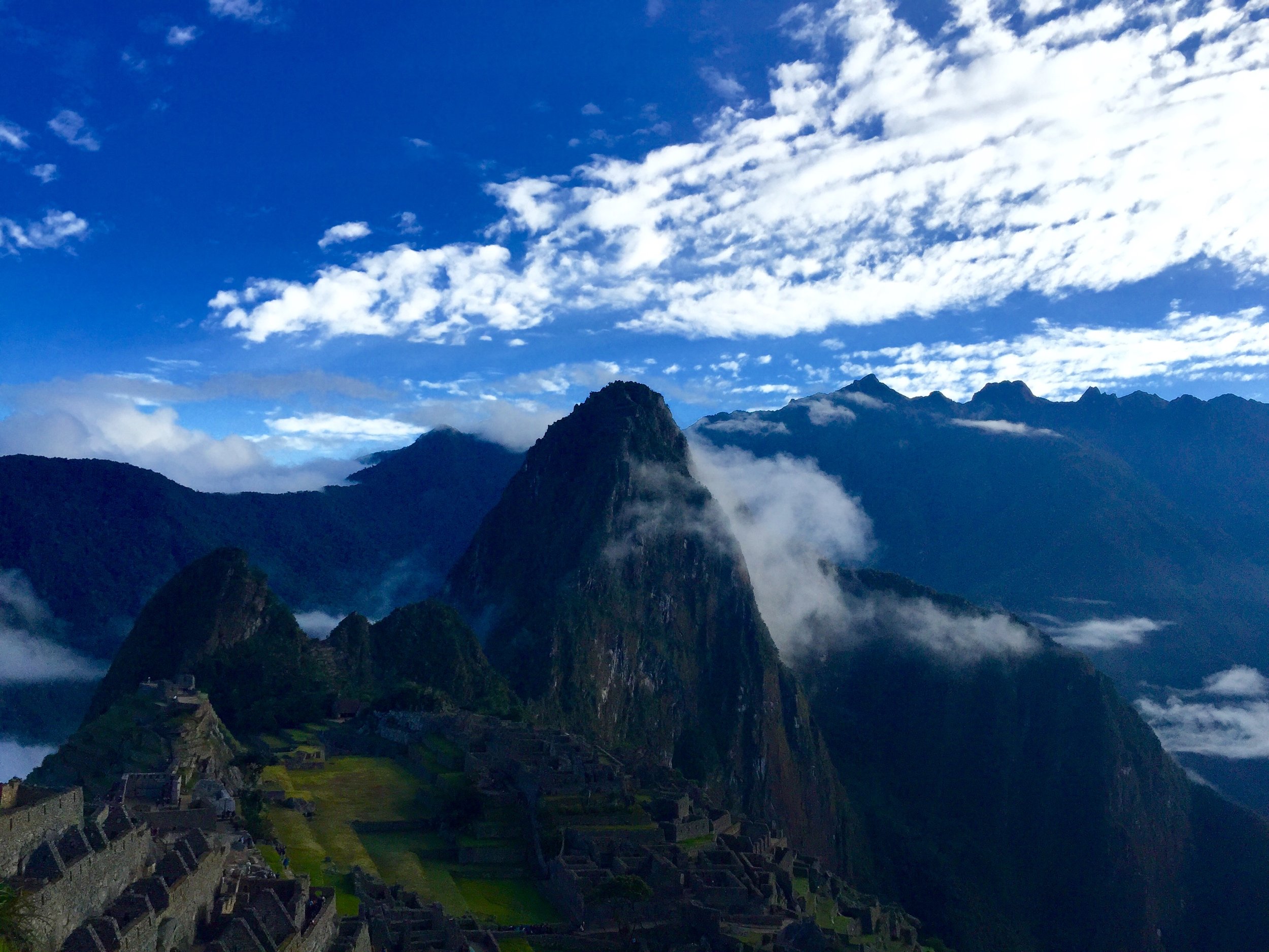 A scenic view of Machu Picchu with ancient stone structures, set against towering green mountains and a bright blue sky with scattered clouds.