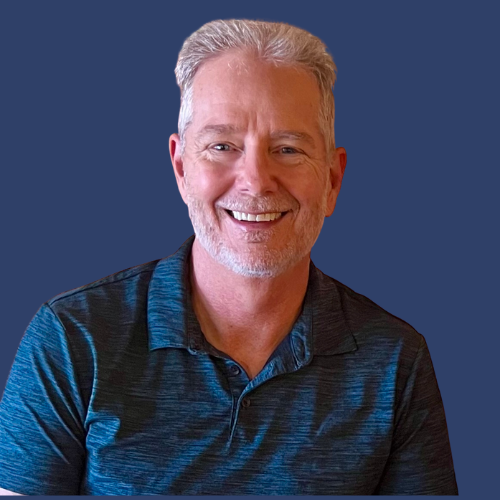 A smiling middle-aged man with gray hair and a beard, wearing a dark blue polo shirt, against a dark background.