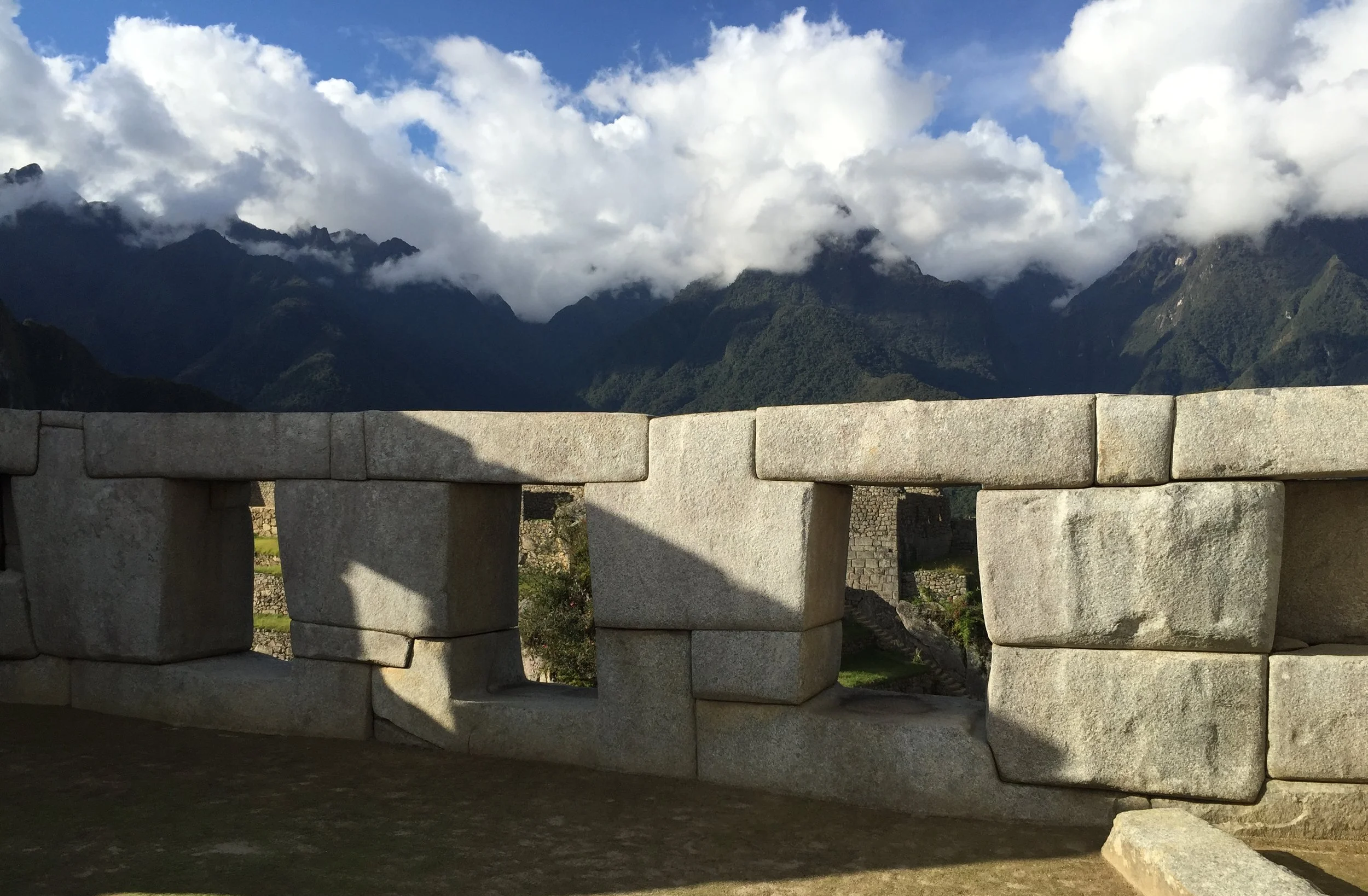 Stone railing with rectangular openings overlooking a mountainous landscape with clouds and green forest.