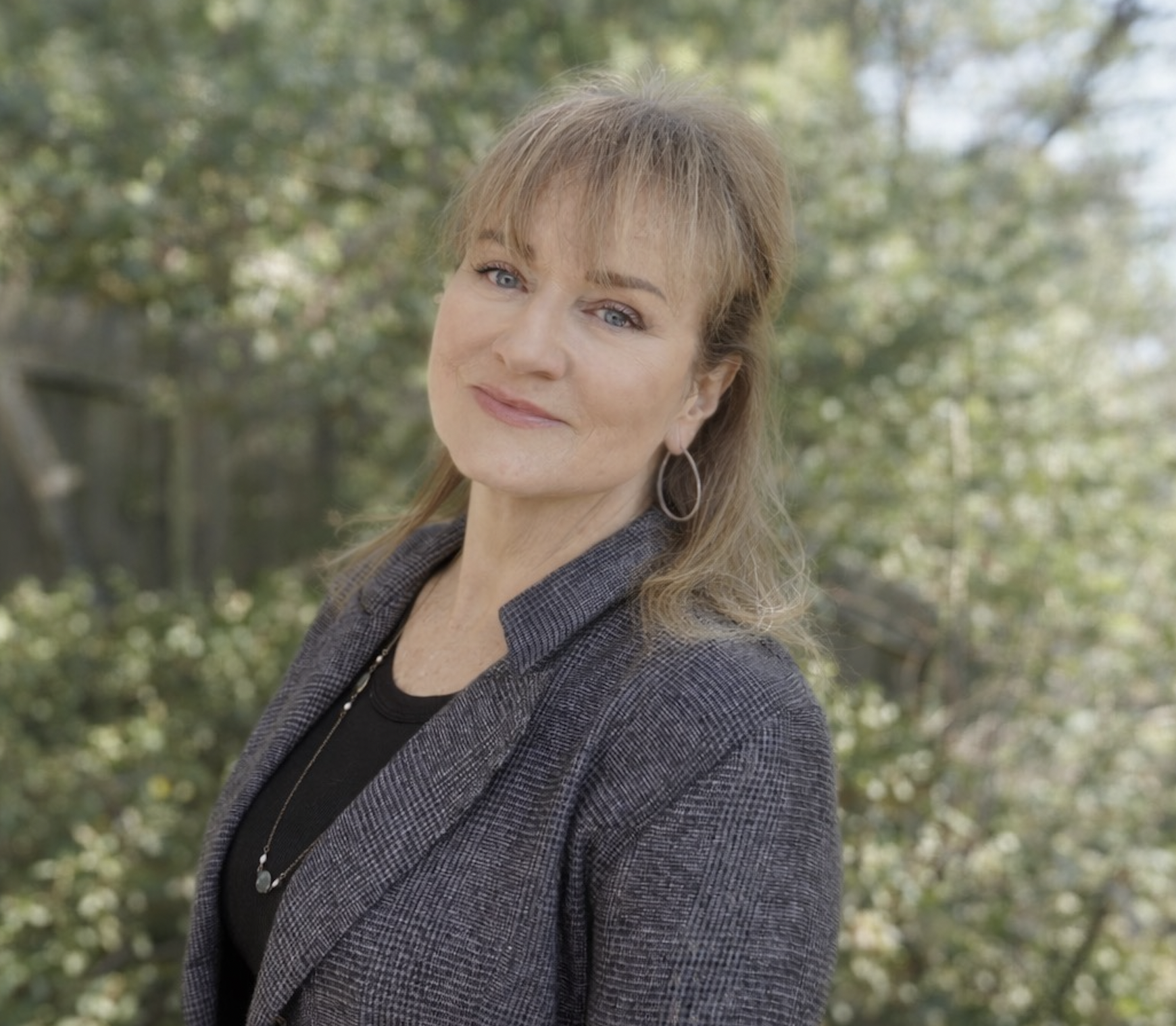 A woman with light brown hair and blue eyes standing outdoors with trees in the background, wearing a dark blazer and a necklace.