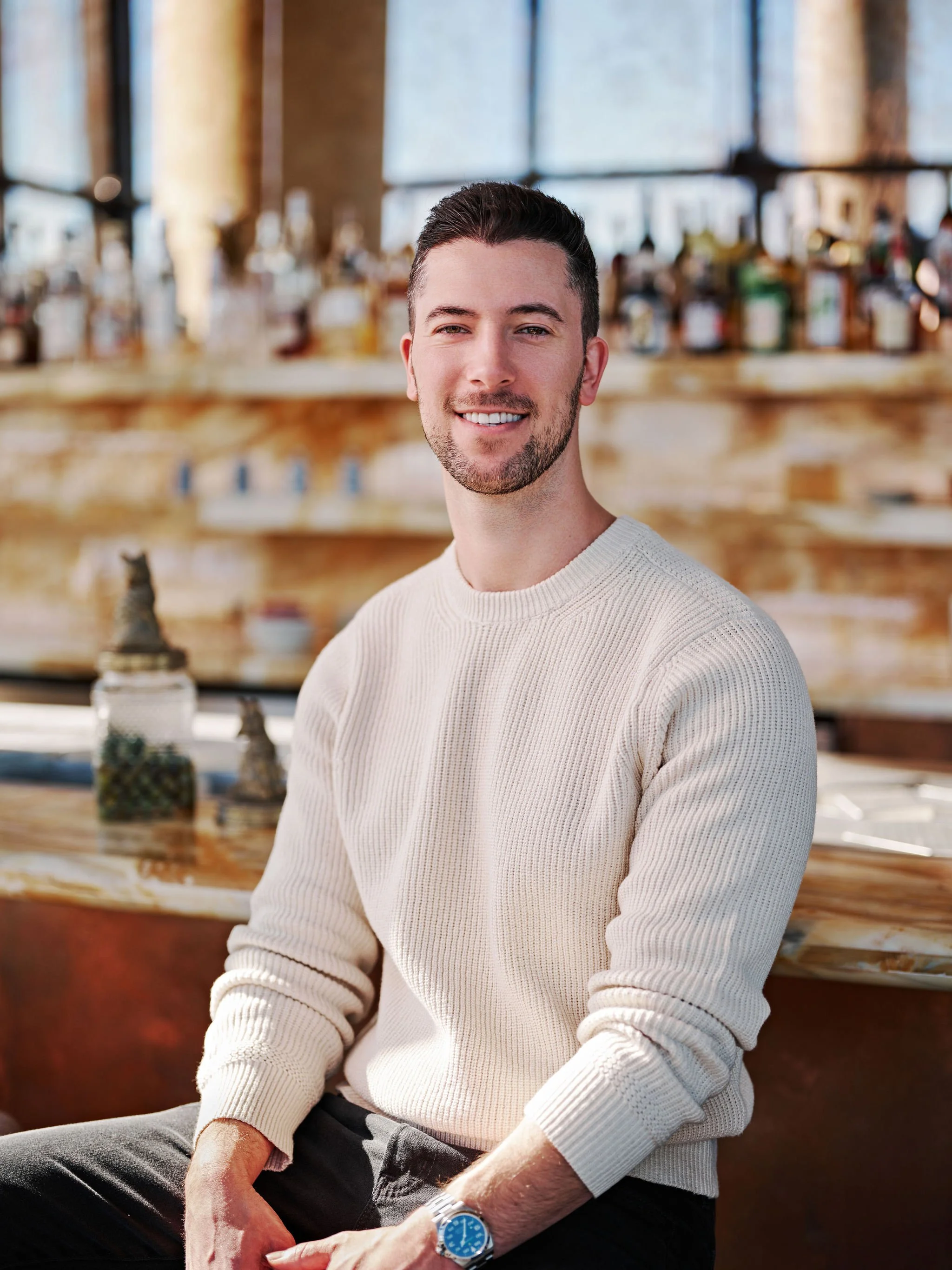 A young man in a navy blue suit, white shirt, and watch sitting on a beige armchair with a wooden wall background, smiling at the camera.