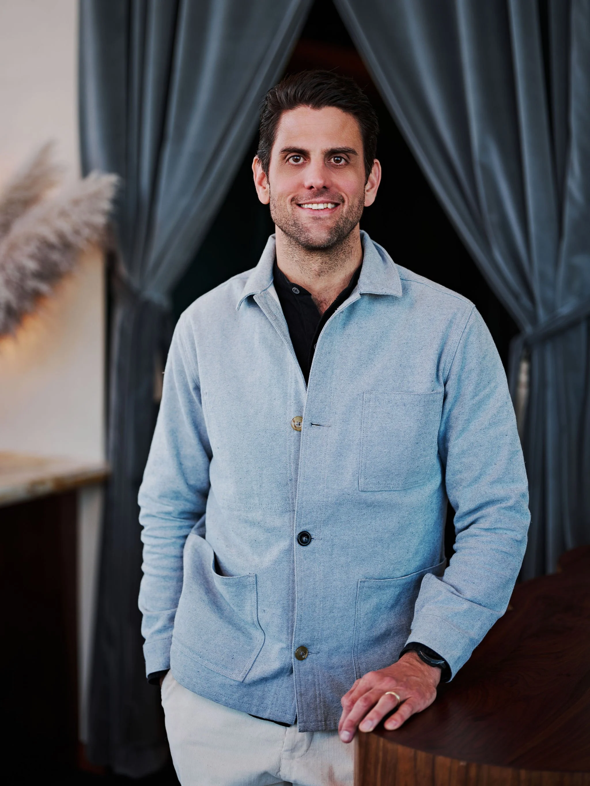 A man standing with arms crossed in a restaurant or cafe, smiling, wearing a colorful striped short-sleeve shirt and light-colored pants, with a drink and a bottle on the counter next to him.