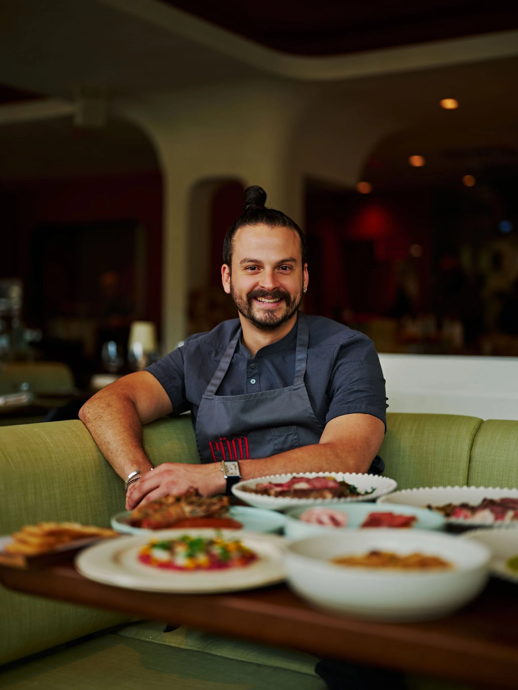 A man with dark hair, beard, and mustache, sitting at a wooden table, smiling, wearing a black shirt and smartwatch, in front of a painted or mural background with a large green leaf.
