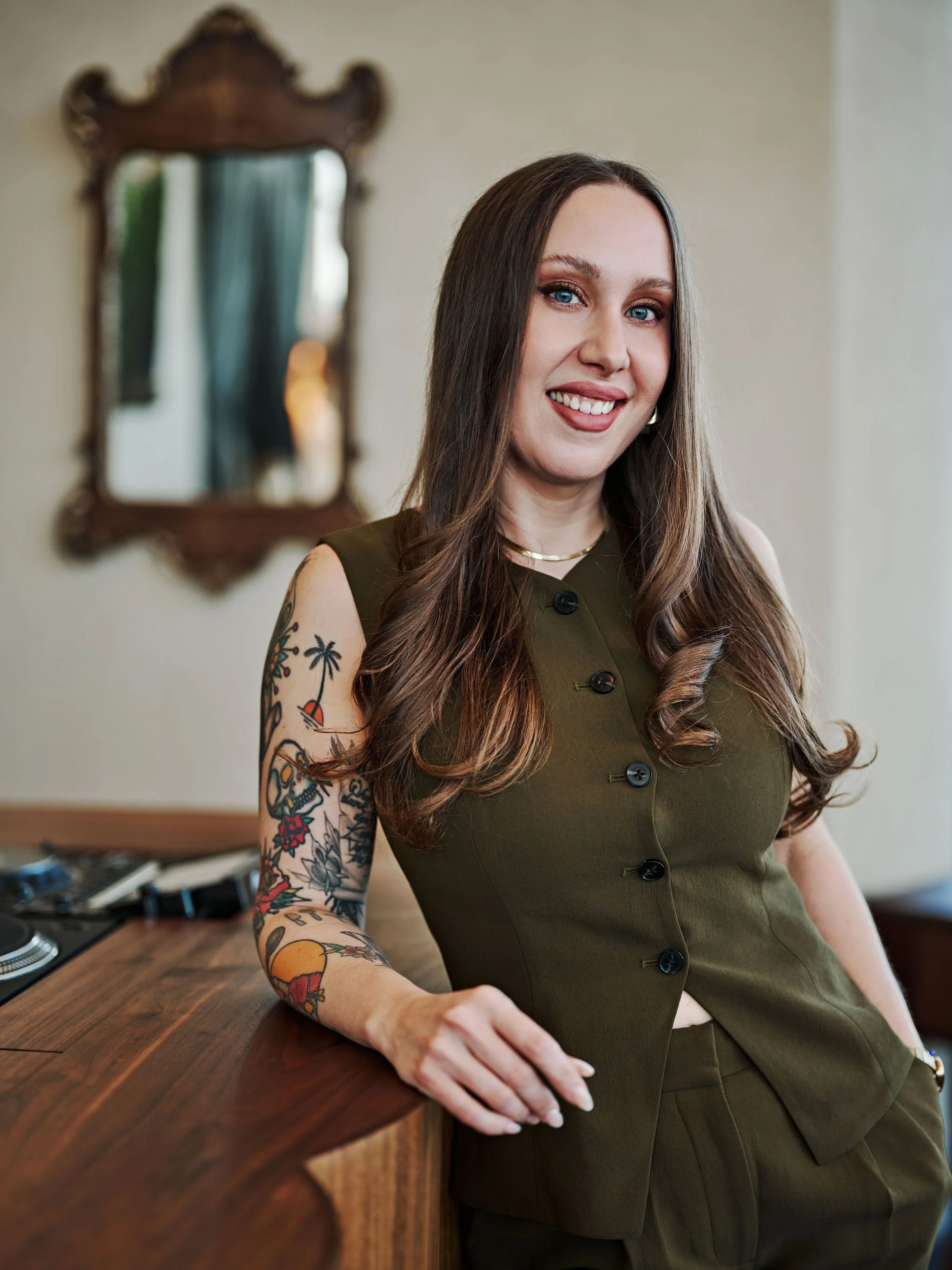 A woman with long brown hair, wearing a white top and a burnt orange blazer, sitting in front of a wooden wall, smiling at the camera.