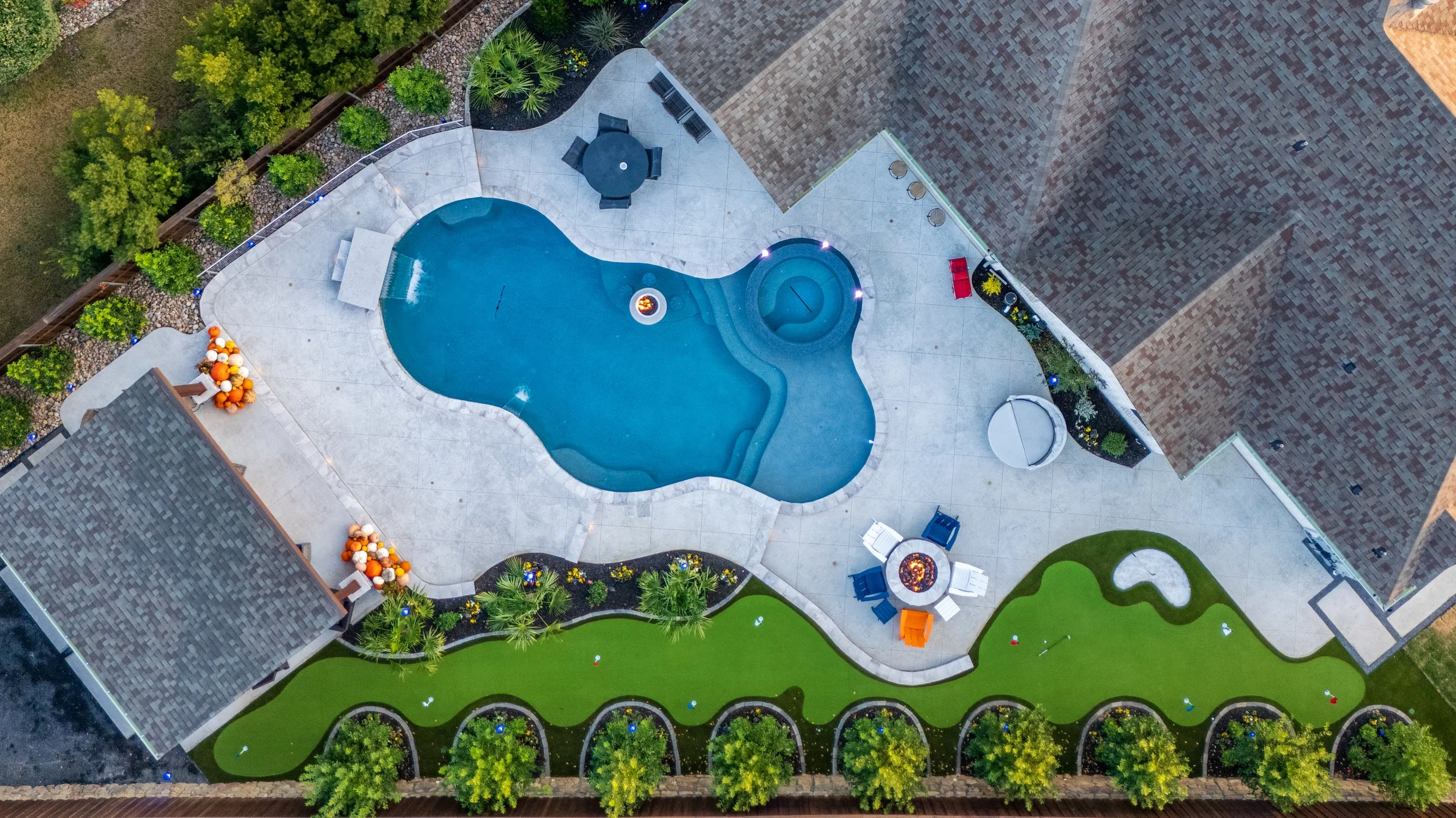 Aerial view of a backyard with a pool, hot tub, outdoor furniture, fire pit, pumpkins, garden, and surrounding landscaping.