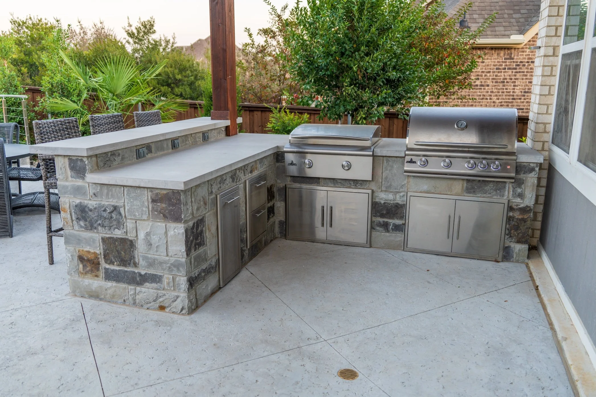 Outdoor kitchen with a stone counter, two stainless steel grills, and storage cabinets, surrounded by patio furniture and greenery.
