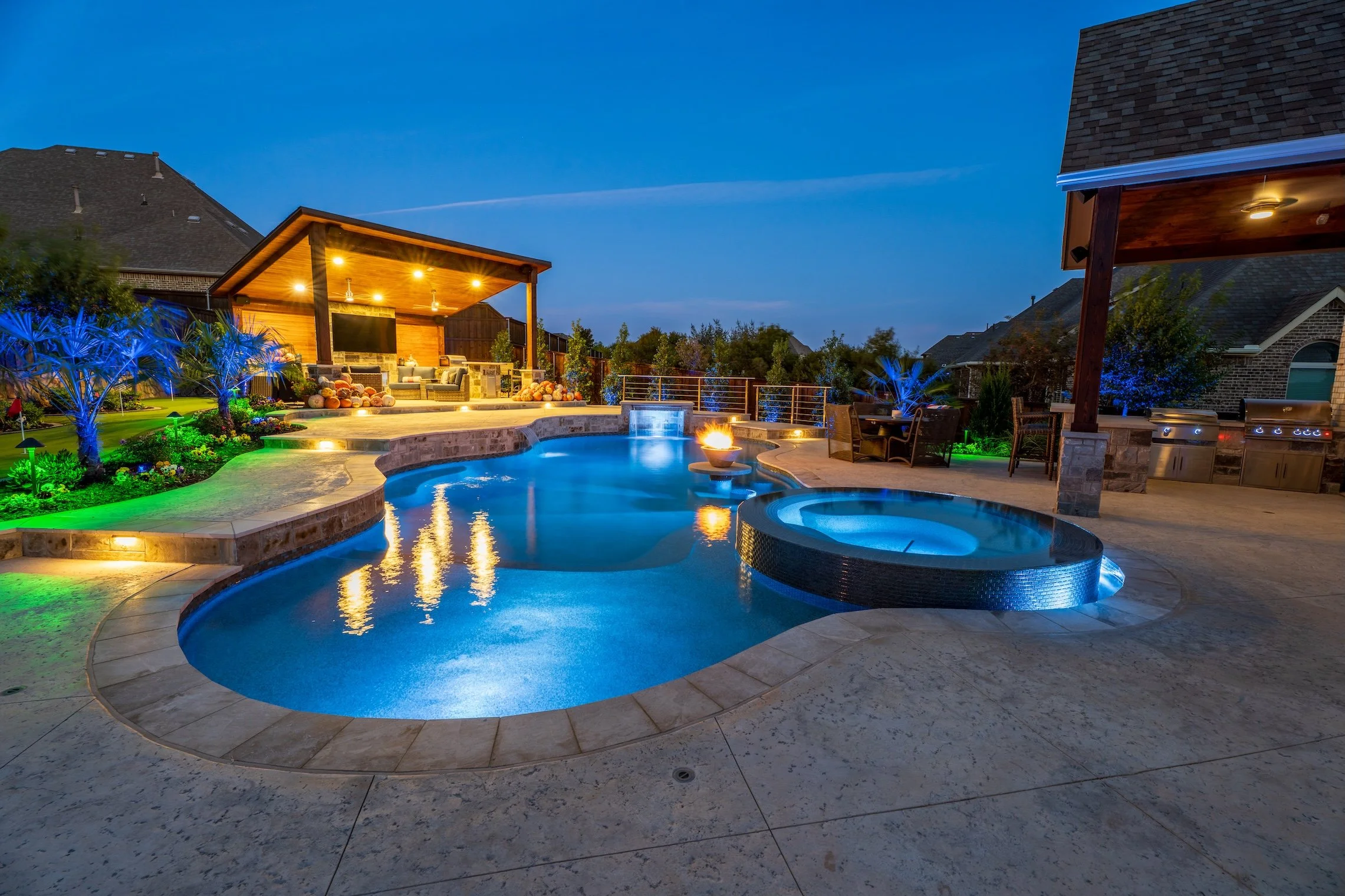 Luxury backyard with illuminated swimming pool, hot tub, fire pit, outdoor seating, landscaping, and outdoor kitchen at dusk.
