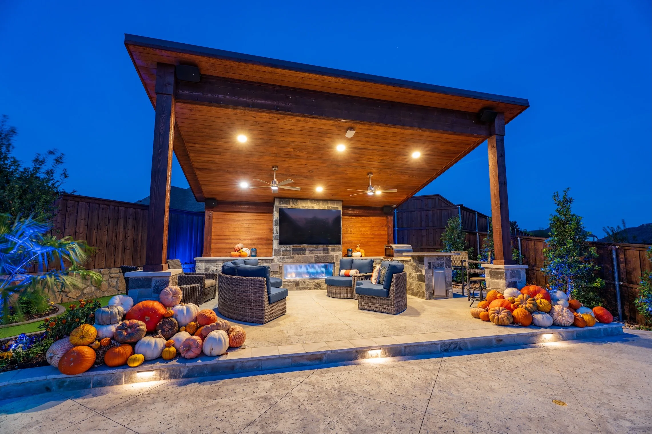 Outdoor covered patio with a stone fireplace, a large flat-screen TV, and seating area with six chairs and cushions. The patio is decorated with pumpkins and gourds, suggesting a fall or Halloween theme. The area is lit with ceiling lights and three 