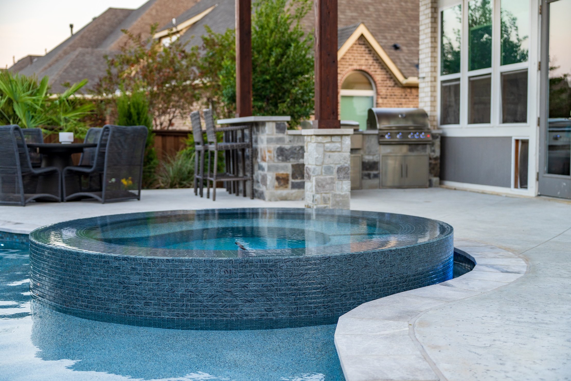 A backyard with a swimming pool and outdoor kitchen area, including a barbecue grill, a bar with stools, and a patio dining set surrounded by greenery.