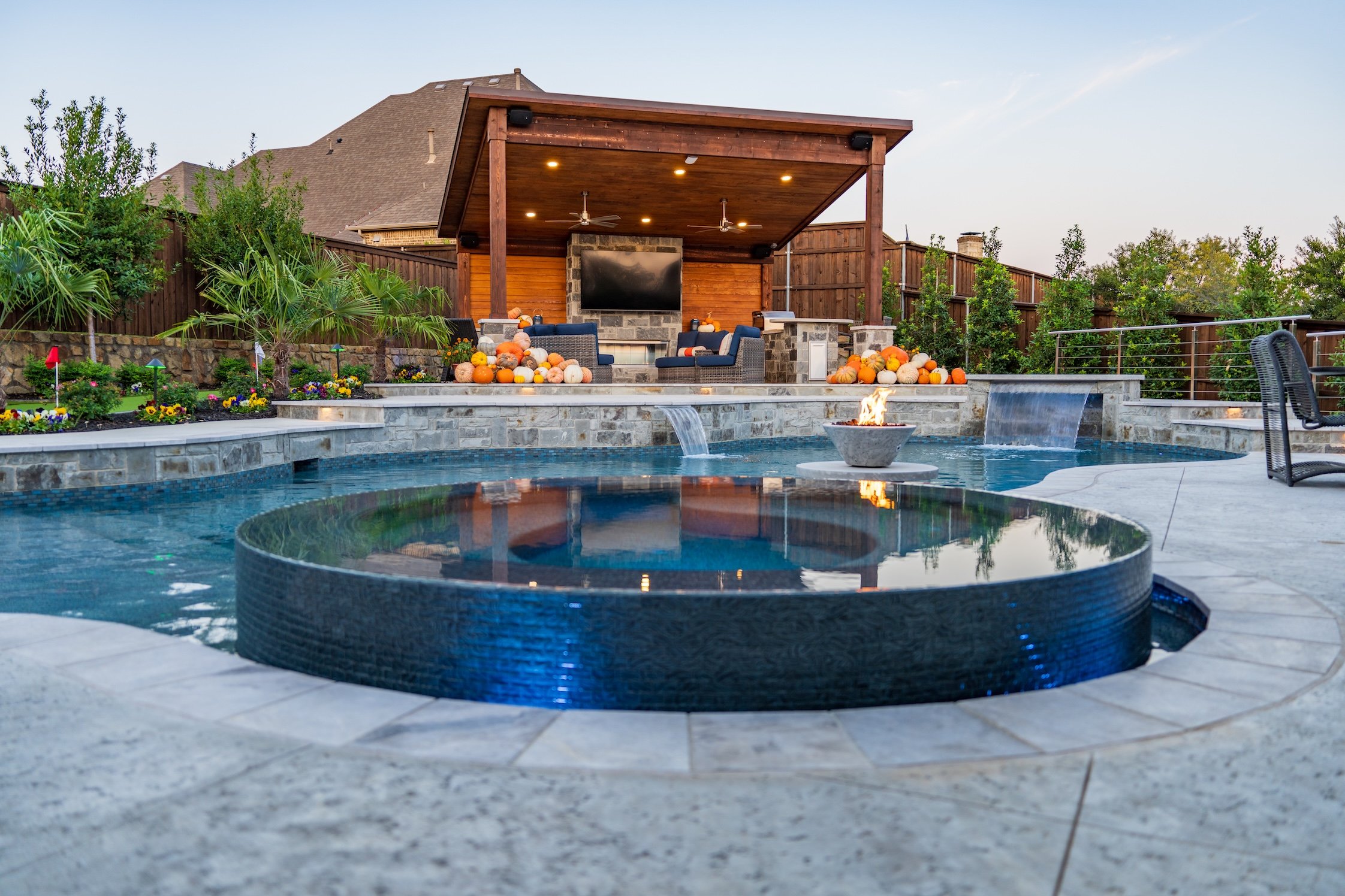 Backyard with a swimming pool, a hot tub, a fire pit, and an outdoor entertainment area with a large TV, surrounded by landscaping and trees.