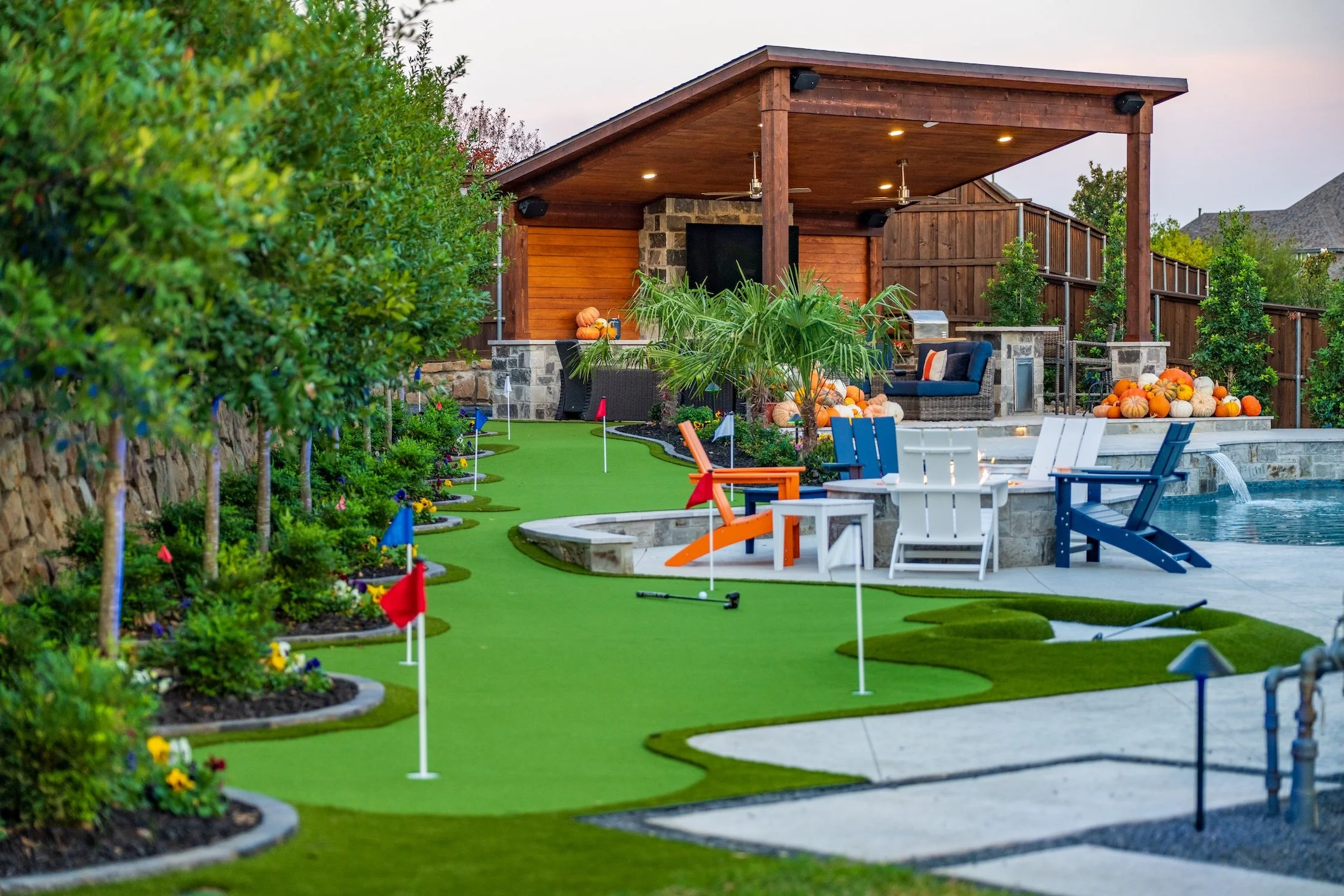 Backyard with a mini-golf putting green, Adirondack chairs, a pool, covered patio with outdoor seating, pumpkins, and landscaped trees and plants.
