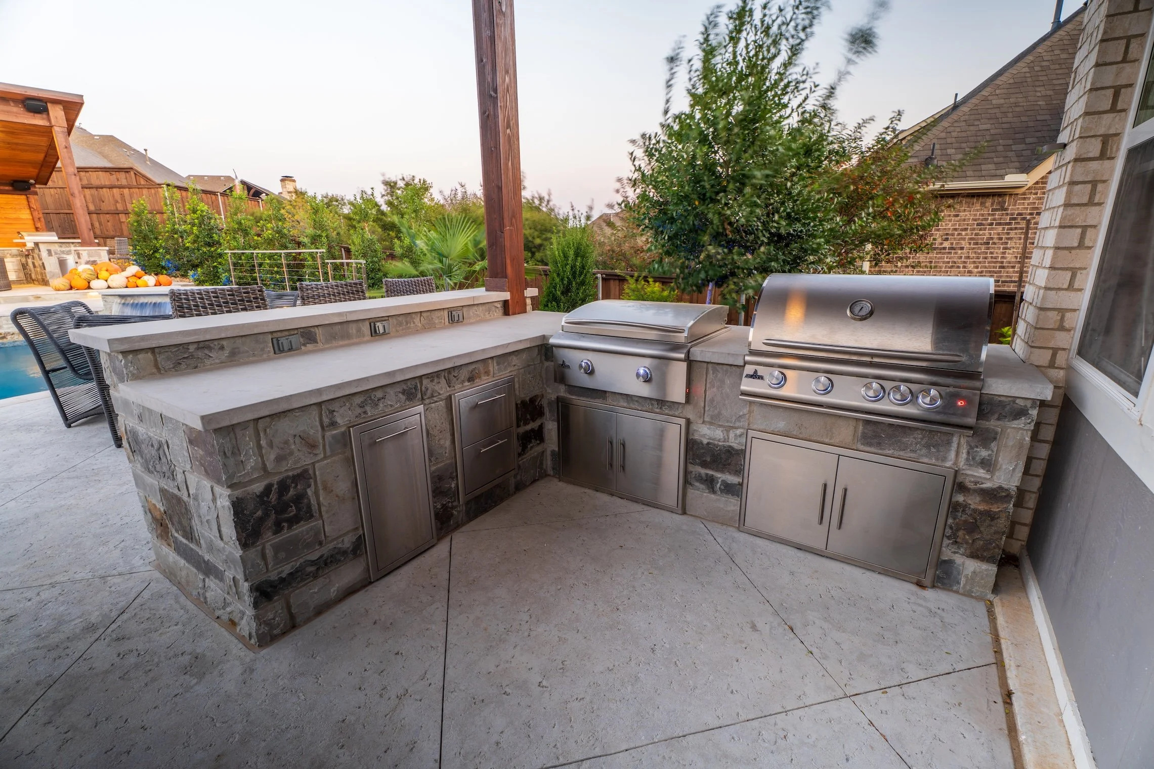 Outdoor kitchen with stone counter, stainless steel grills, and cabinet storage, nearby a backyard with trees and lounge chairs by a pool.