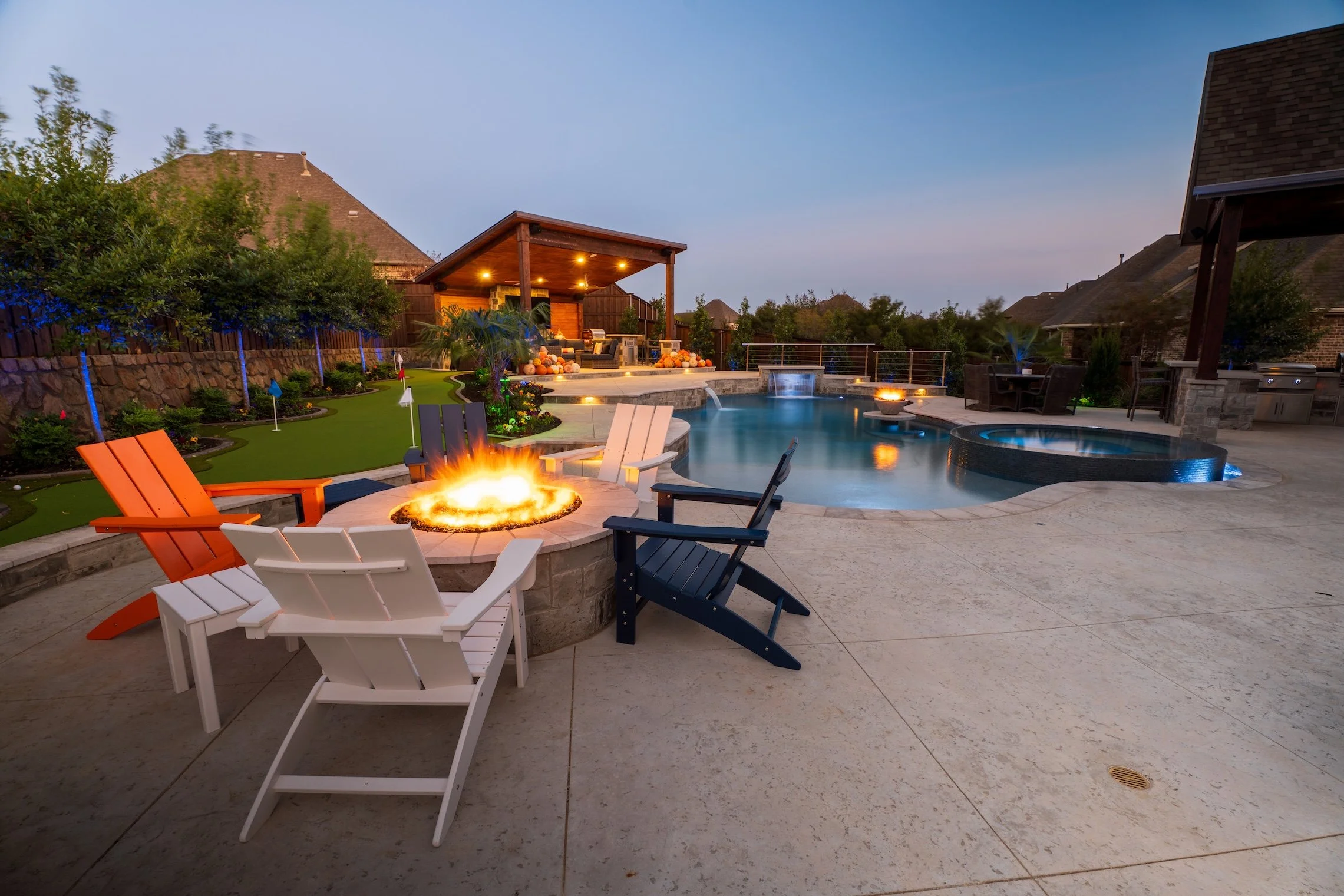 Backyard pool area with fire pit, pool, hot tub, outdoor seating, and landscaped yard at dusk