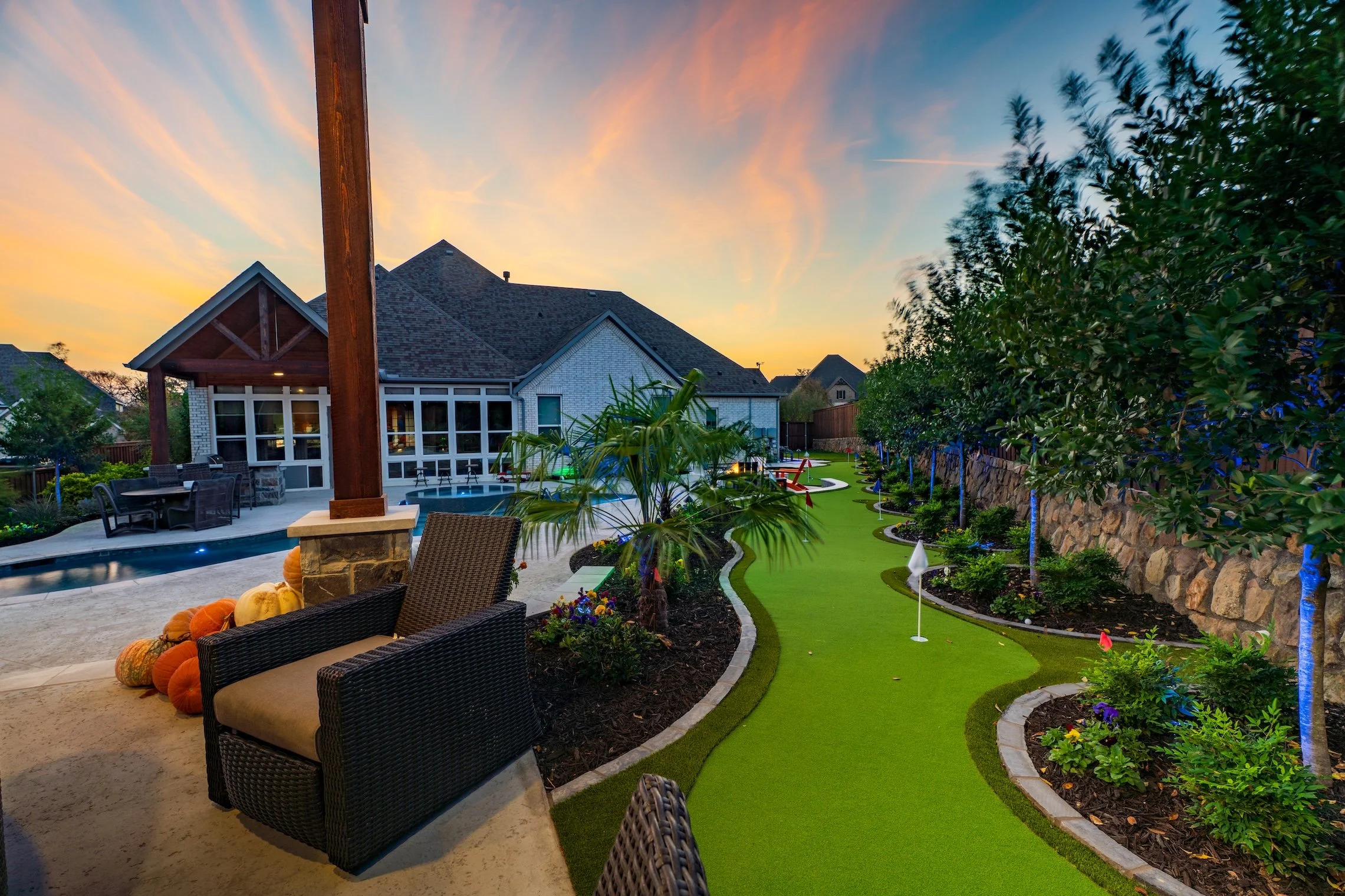 Backyard with a putting green, a patio with a table and chairs, a hot tub, and a house with large windows. The scene is during sunset with colorful sky and landscaping including trees, shrubs, and pumpkins.