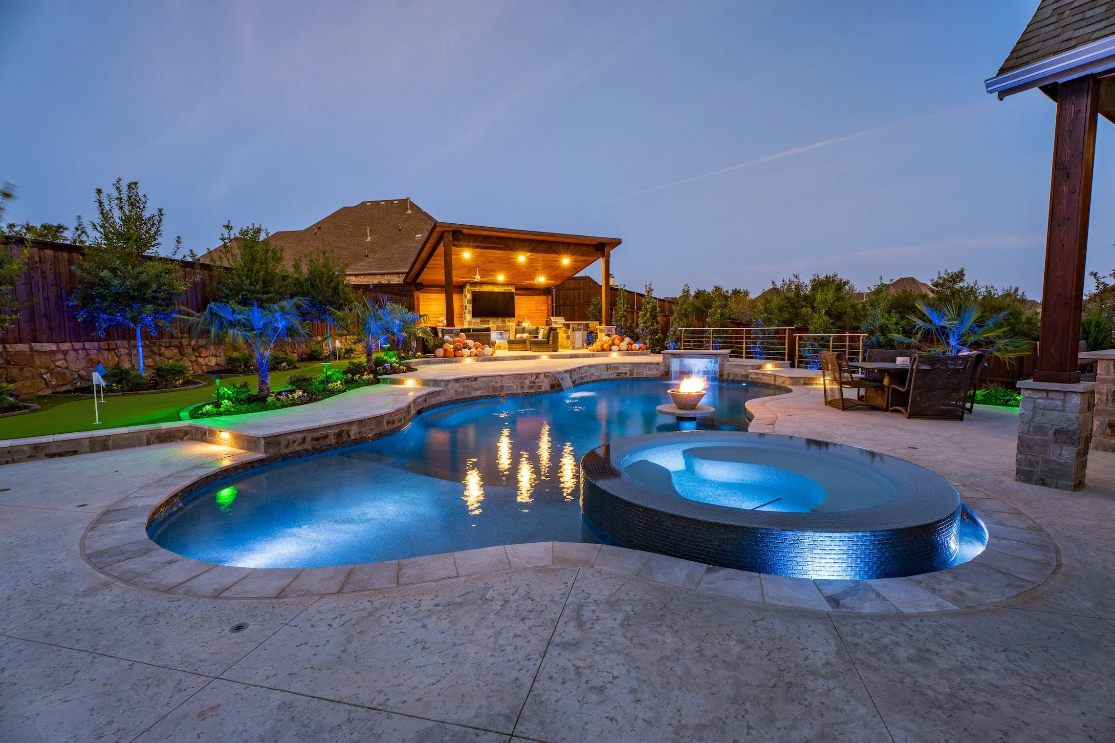Luxury backyard with a swimming pool, hot tub, fire pit, outdoor seating area, and landscaped garden under evening sky.